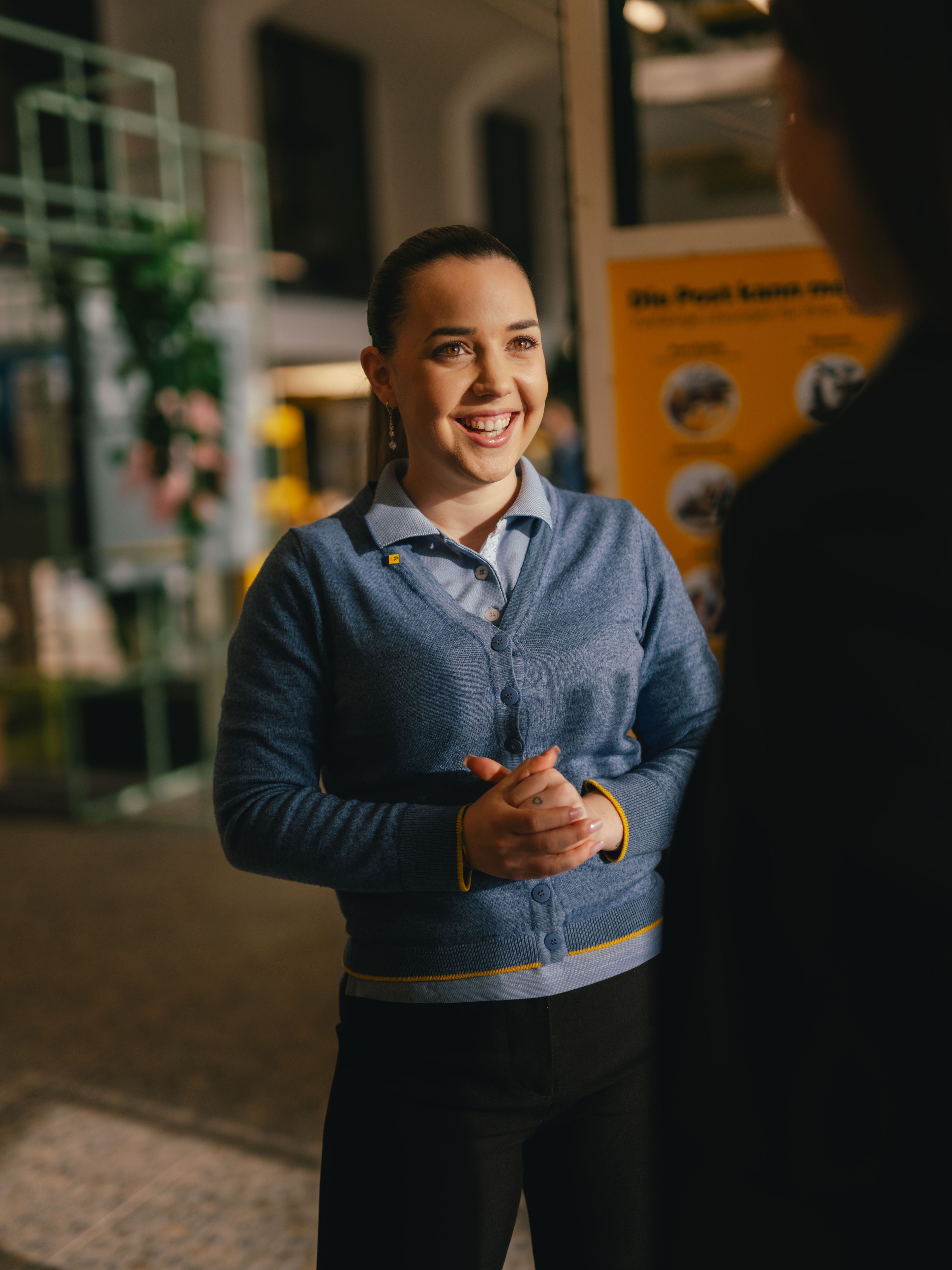 Eine Person steht in einem Innenraum und spricht mit einer weiteren Person im Vordergrund. Die Person trägt eine blaue Strickjacke über einem Hemd und hält die Hände vor dem Körper zusammen. Im Hintergrund befinden sich Infotafeln und Ausstellungsstrukturen.