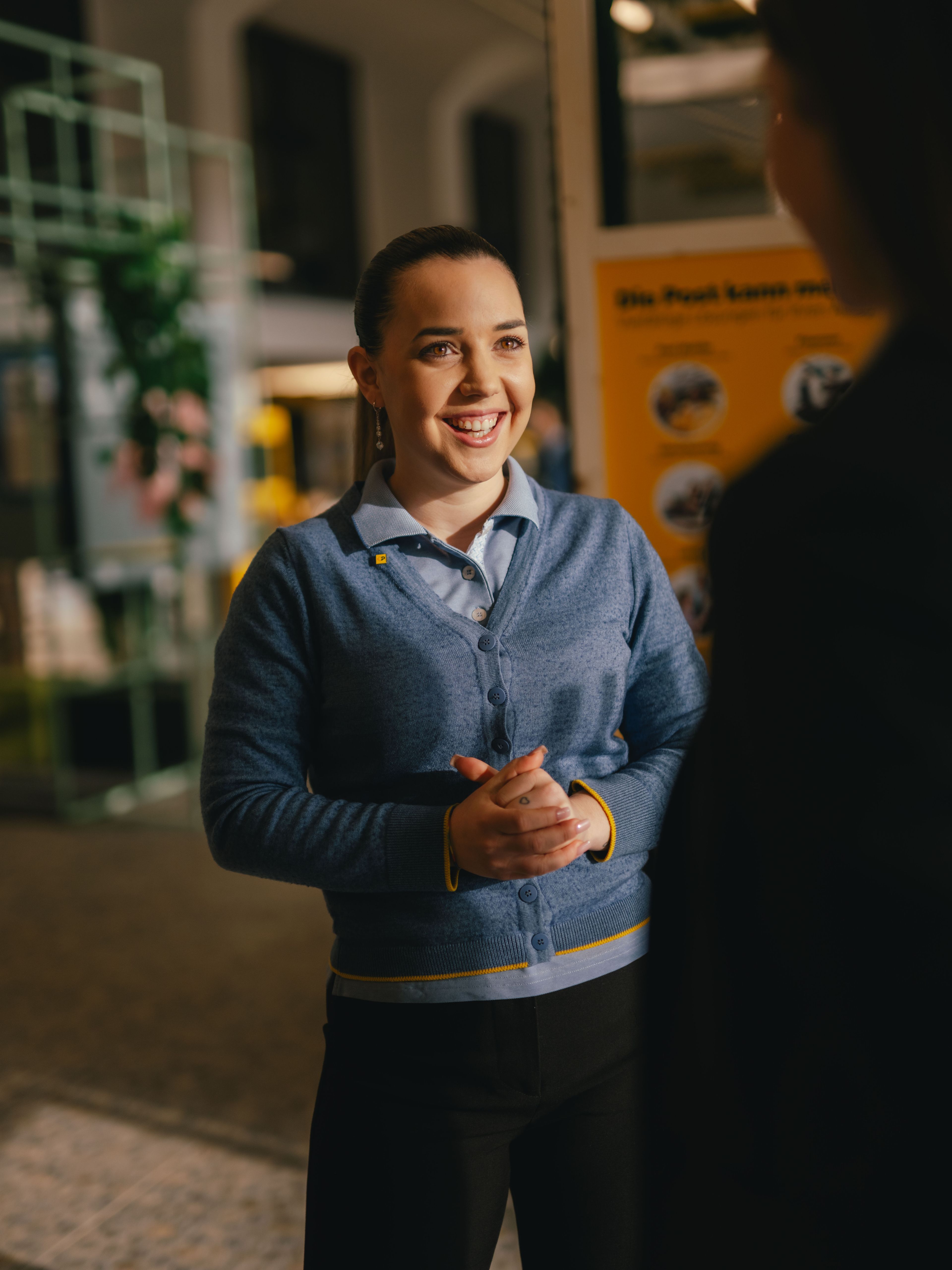 Une personne se tient à l’intérieur et s’adresse à une autre personne au premier plan. La personne porte un cardigan bleu par-dessus une chemise et garde les mains jointes devant elle. À l’arrière-plan se trouvent des panneaux d’information et des structures d’exposition.