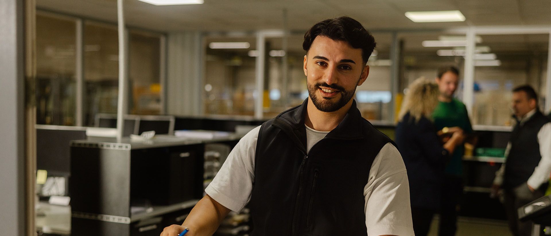 Ein Zollbeamter stempelt Dokumente an einem Schalter, im Hintergrund ein Büro mit mehreren Personen.