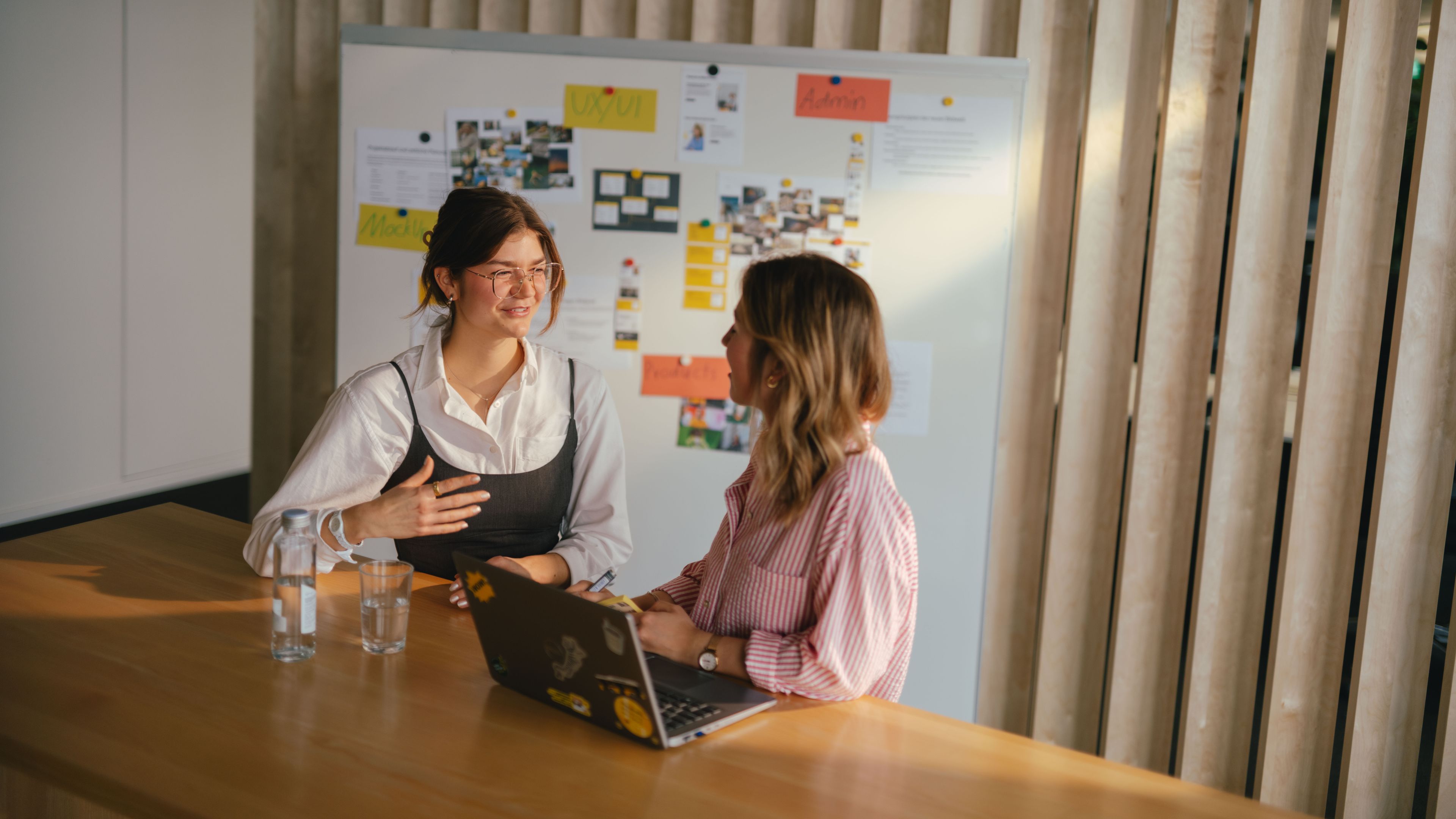 Zwei Personen sitzen an einem langen Holztisch in einem modernen Innenraum. Vor ihnen steht ein aufgeklappter Laptop mit mehreren Aufklebern darauf sowie zwei Gläser Wasser. Im Hintergrund befindet sich eine grosse Pinnwand mit vielfältigen Notizzetteln, Fotos und farbigen Karten. Hinter der Pinnwand sind vertikal angeordnete Holzlamellen sichtbar, durch die warmes Licht in den Raum fällt.