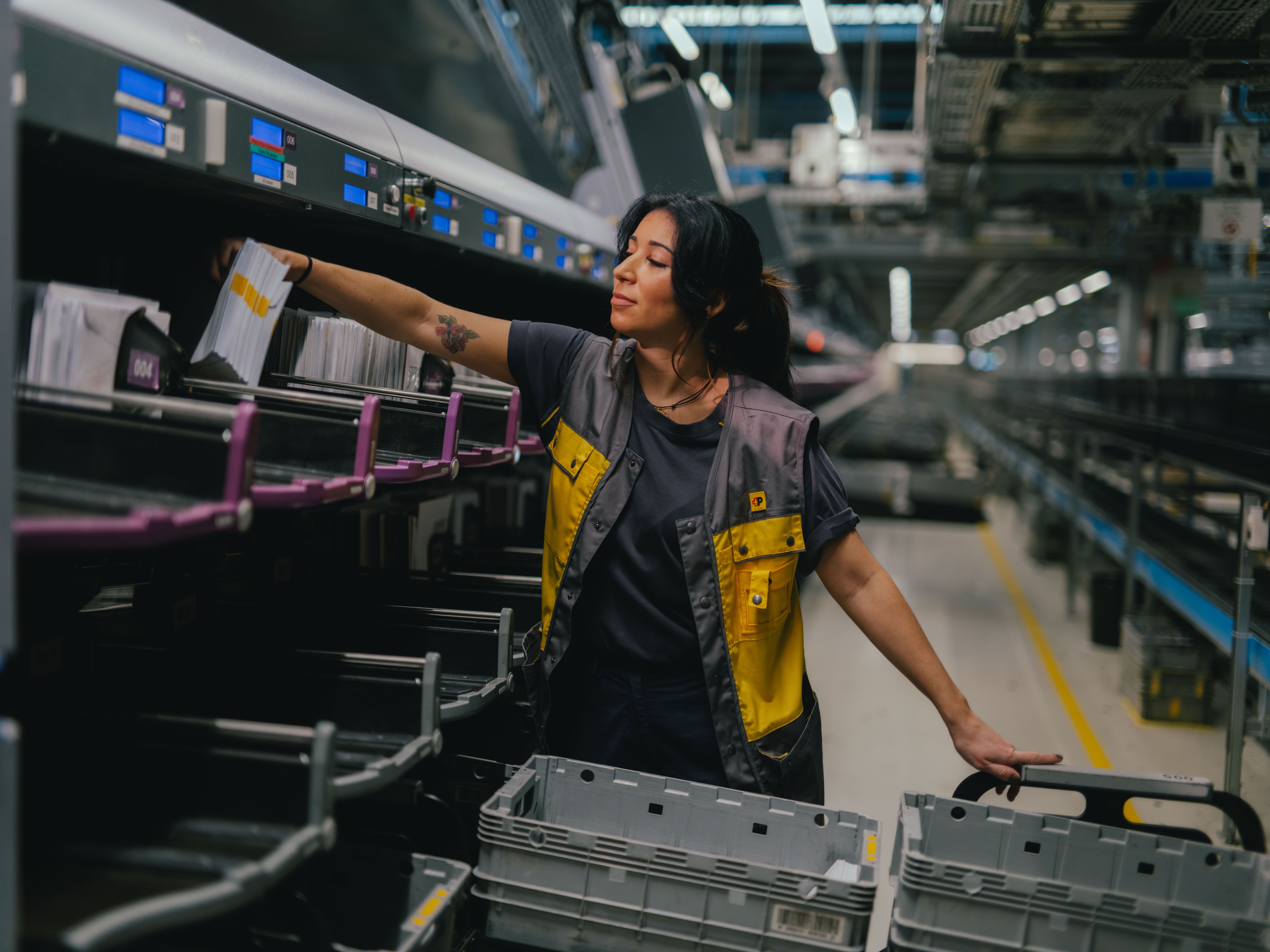 A person is standing in a large, brightly lit hall and sorting mail items into an automated compartment system. The person is wearing grey and yellow work clothing and is reaching into one of the sorting slots. Several grey plastic bins are positioned in front. In the background, machinery, conveyor systems, and industrial structures are visible.