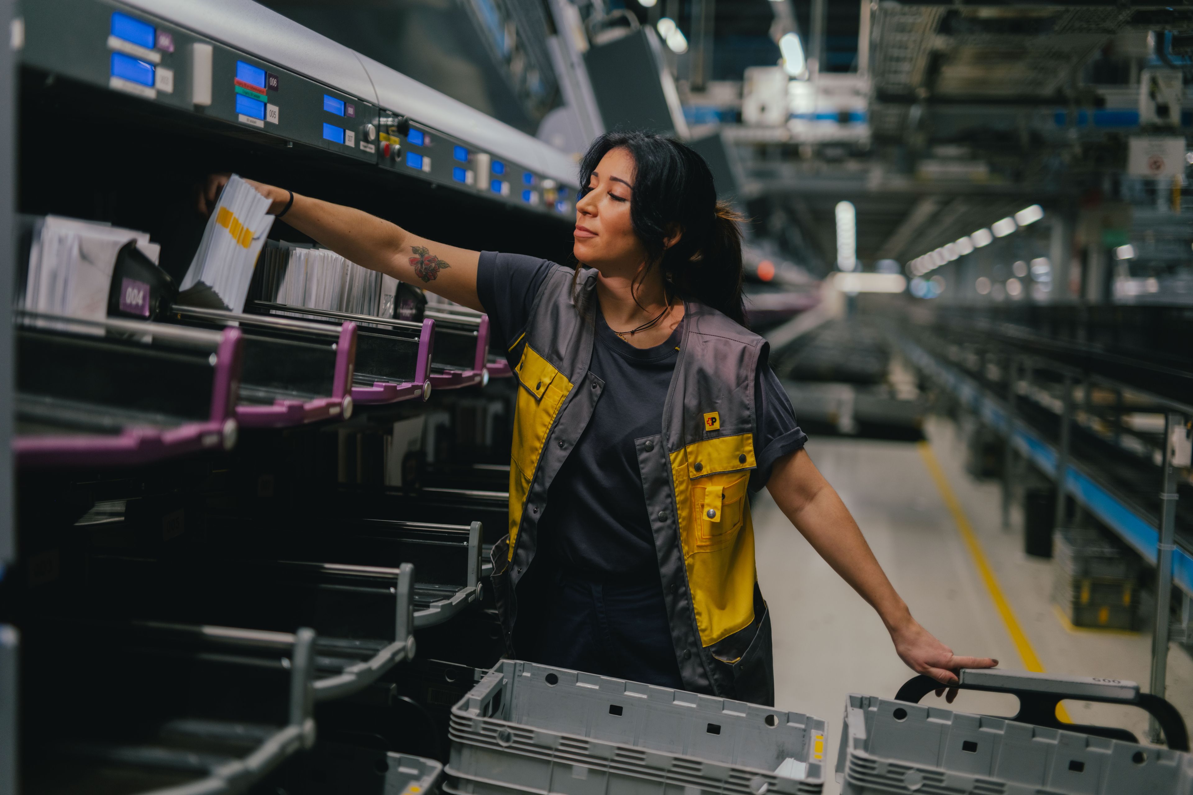 A person is standing in a large, brightly lit hall and sorting mail items into an automated compartment system. The person is wearing grey and yellow work clothing and is reaching into one of the sorting slots. Several grey plastic bins are positioned in front. In the background, machinery, conveyor systems, and industrial structures are visible.