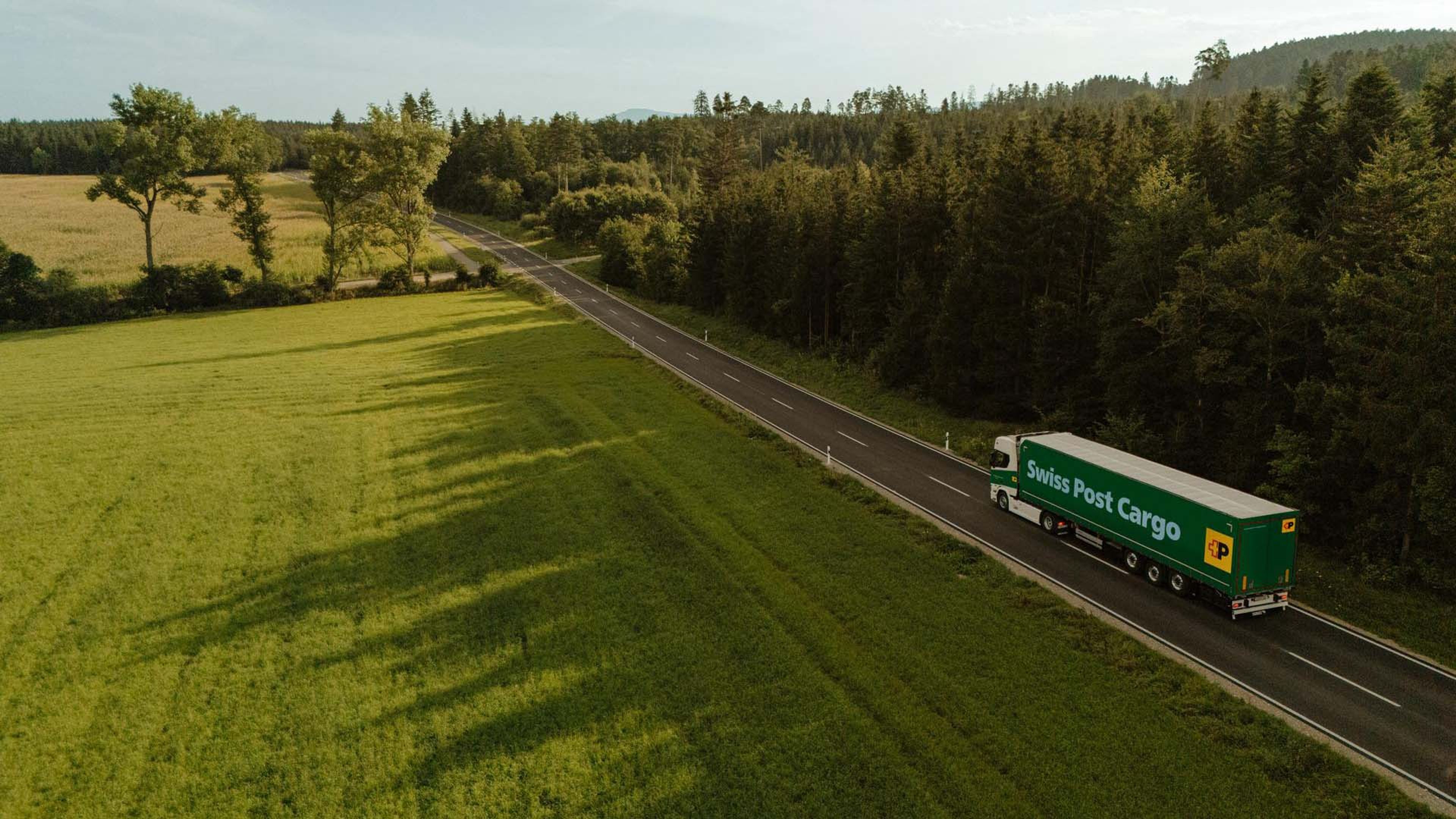 Swiss Post Cargo lorry driving on a country road between a meadow and a forest.