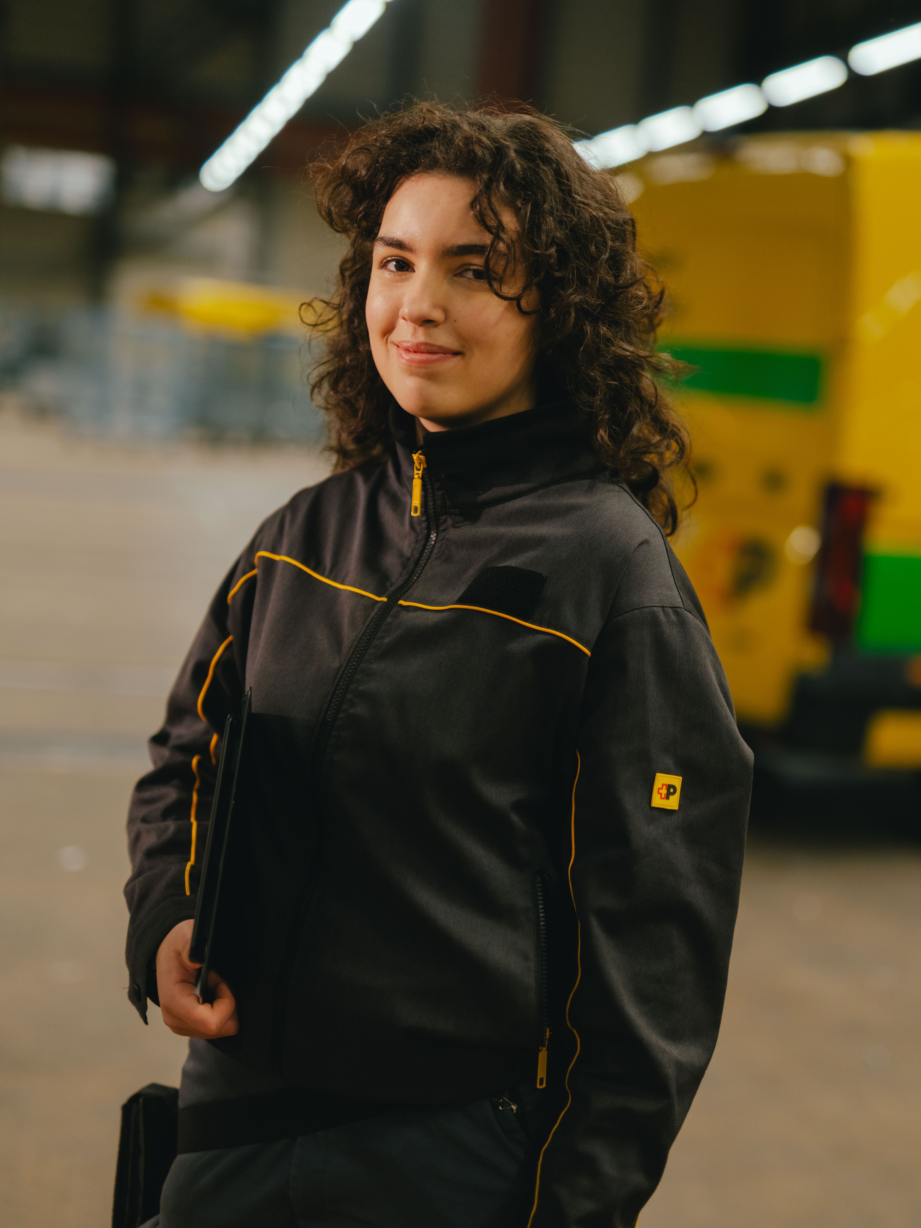 Eine Person steht in einer grossen Halle und trägt dunkelgraue Arbeitskleidung mit gelben Akzenten. Die Person hält ein dunkles Tablet oder Klemmbrett am Körper. Im Hintergrund befindet sich ein gelb‑grünes Lieferfahrzeug mit schwarzer Beschriftung, sowie industrielle Strukturen und hohe Hallendecken.