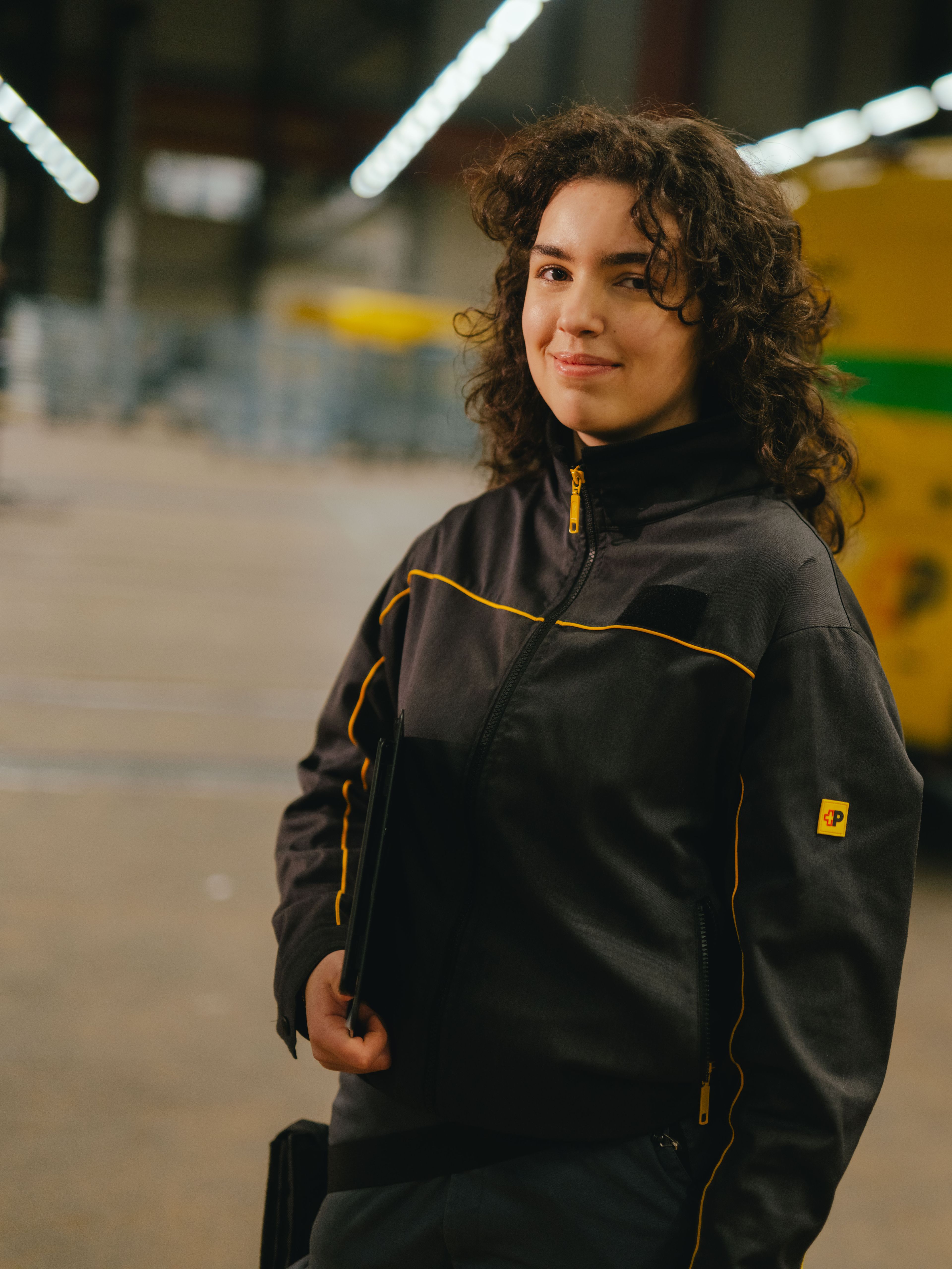 Eine Person in dunkler Arbeitskleidung mit gelben Akzenten steht in einer grossen Halle. Die Person hält einen Gegenstand in der Hand. Im Hintergrund befindet sich ein gelb‑grünes Lieferfahrzeug mit schwarzer und roter Beschriftung sowie eine weitläufige, gut beleuchtete Lagerfläche.