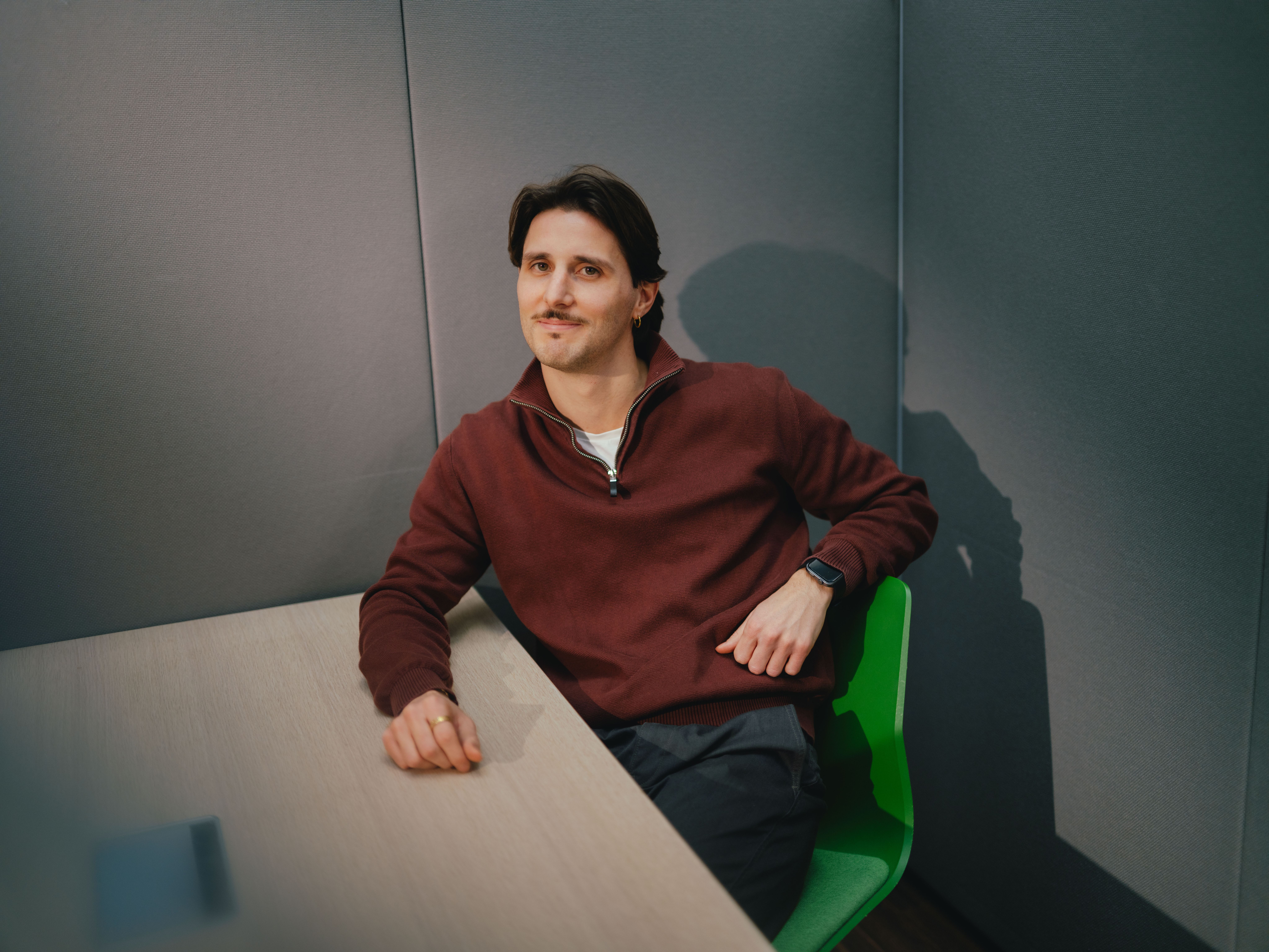 Une personne est assise dans un angle de pièce à une table claire. La personne porte un pull rouge foncé et un pantalon sombre et est assise sur une chaise verte. Un bras repose sur la table, l’autre est appuyé sur le dossier de la chaise. L’arrière‑plan est composé de panneaux muraux gris rembourrés.