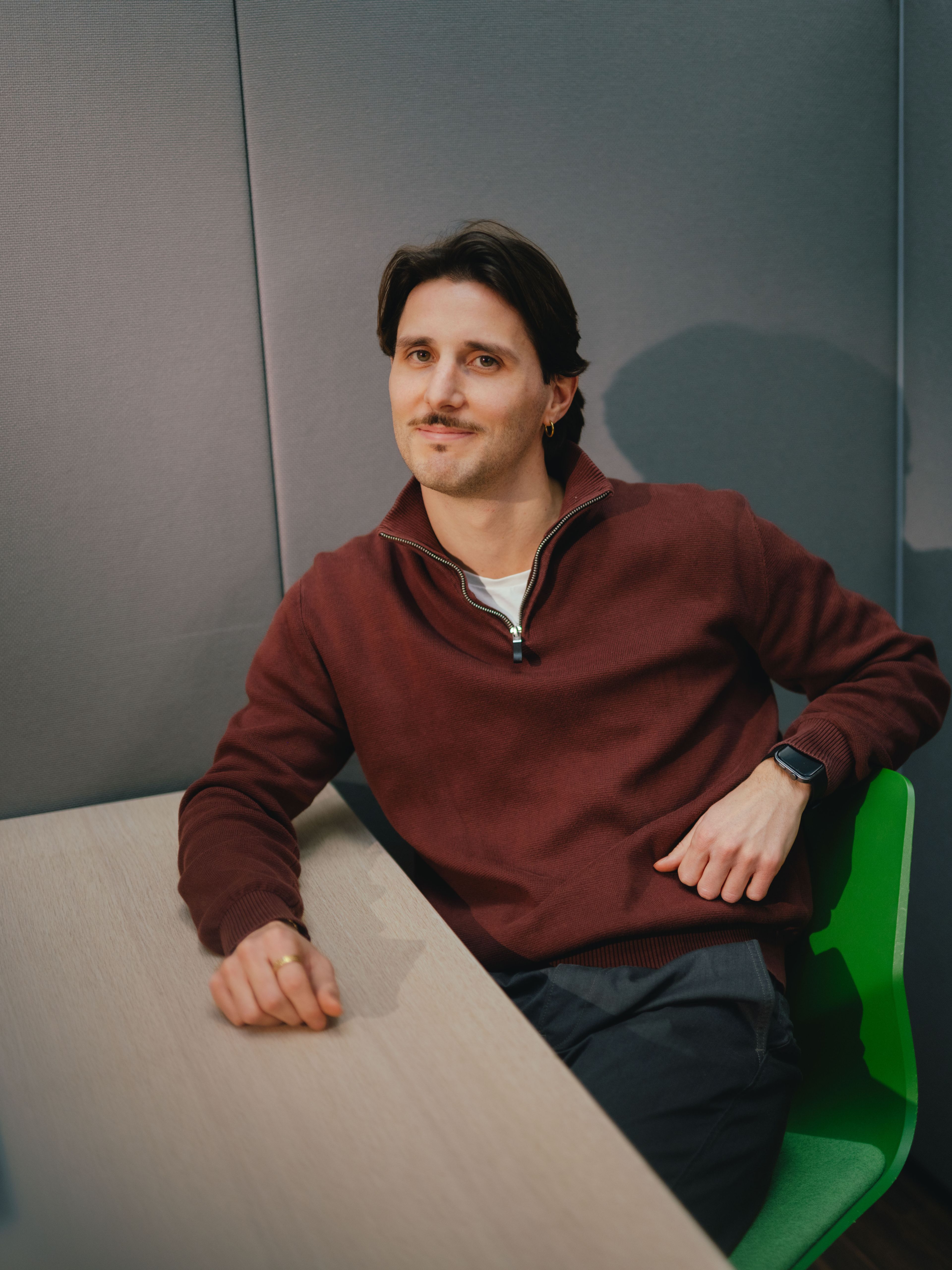 Une personne est assise dans un angle de pièce à une table claire. La personne porte un pull rouge foncé et un pantalon sombre et est assise sur une chaise verte. Un bras repose sur la table, l’autre est appuyé sur le dossier de la chaise. L’arrière‑plan est composé de panneaux muraux gris rembourrés.