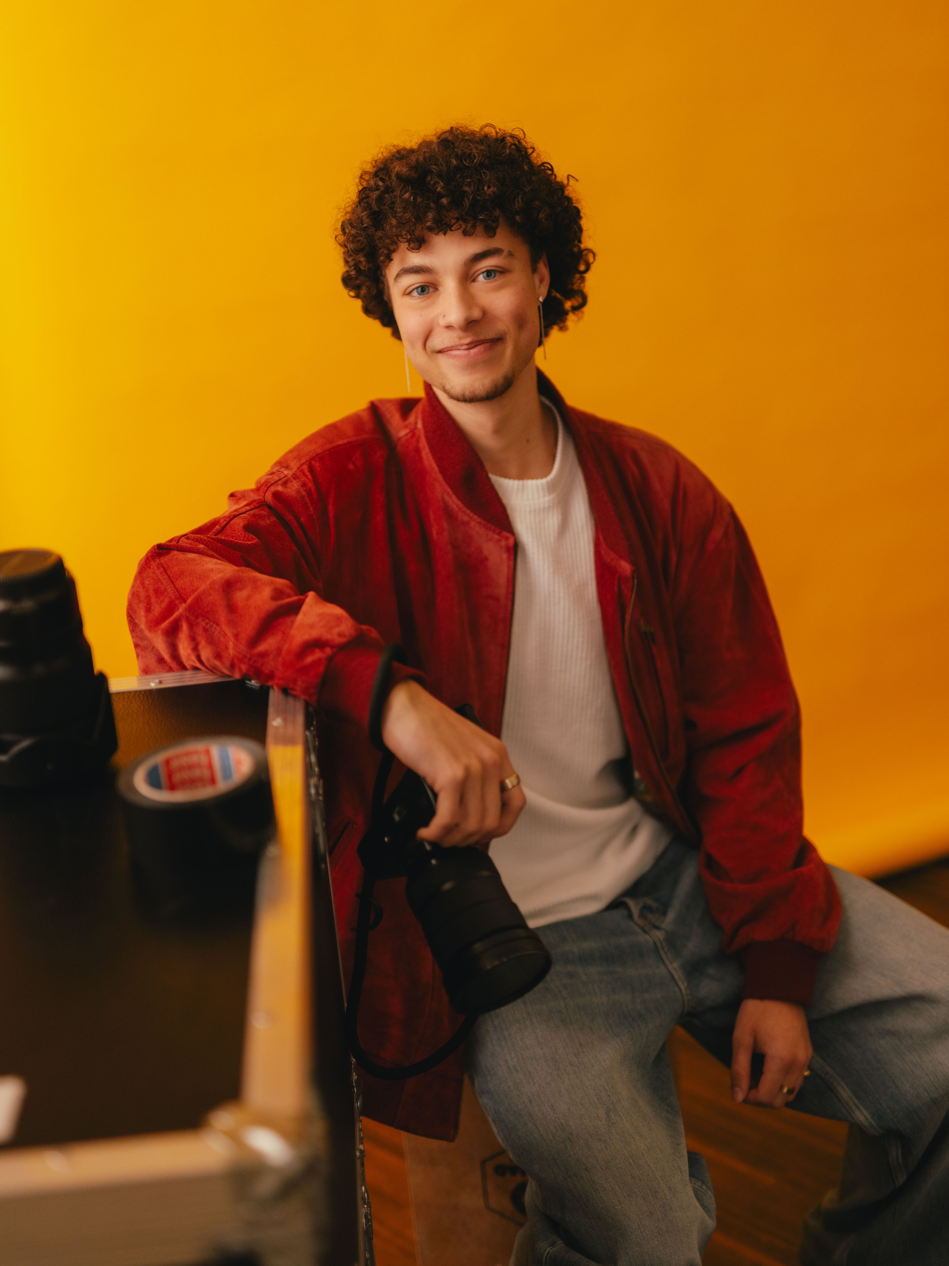 Eine Person sitzt vor einem leuchtend gelben Hintergrund auf einem Transportkoffer. Auf dem Koffer stehen Kameraobjektive und eine Rolle Klebeband. Die Person trägt eine rote Jacke, ein weisses T‑Shirt und eine locker sitzende Jeans und hält eine Kamera locker in einer Hand. Der Raum wirkt wie ein Fotostudio mit professioneller Ausrüstung.