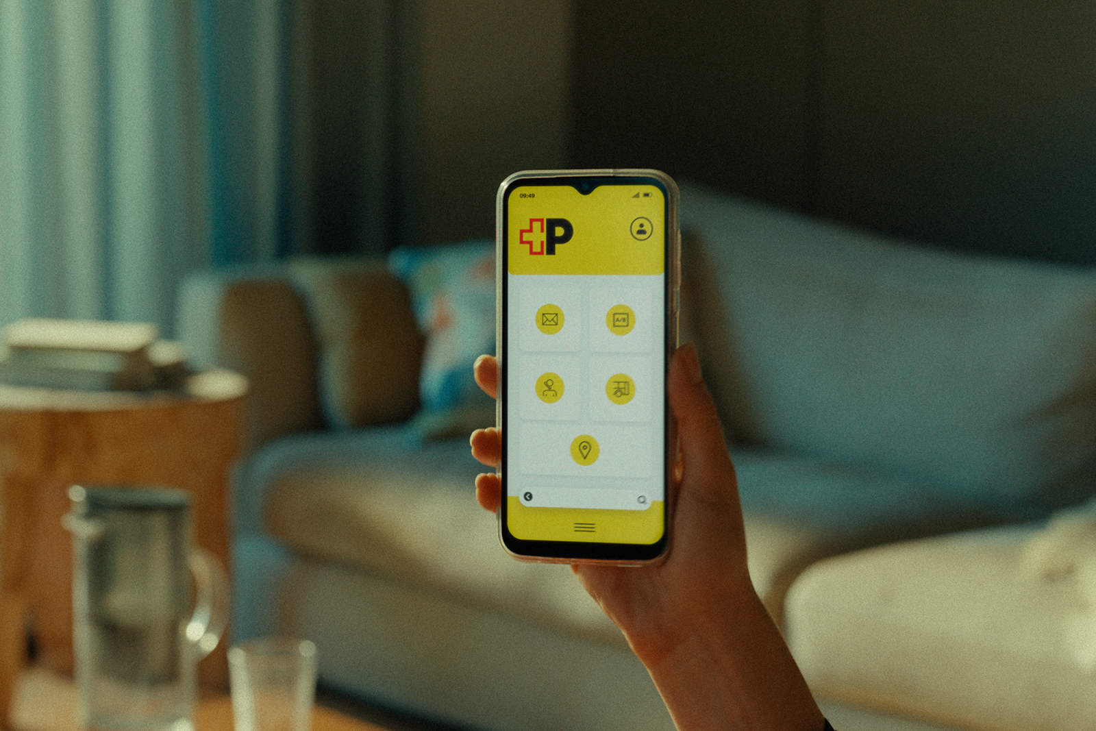Eine Hand hält ein Smartphone vor einem Sofa in einem Wohnzimmer. Auf dem Smartphone‑Bildschirm ist eine gelbe Benutzeroberfläche mit dem rot‑schwarzen Post‑Logo und mehreren runden Symbolen zu sehen. Im Hintergrund stehen ein Sofa, ein Tisch und Haushaltsgegenstände.