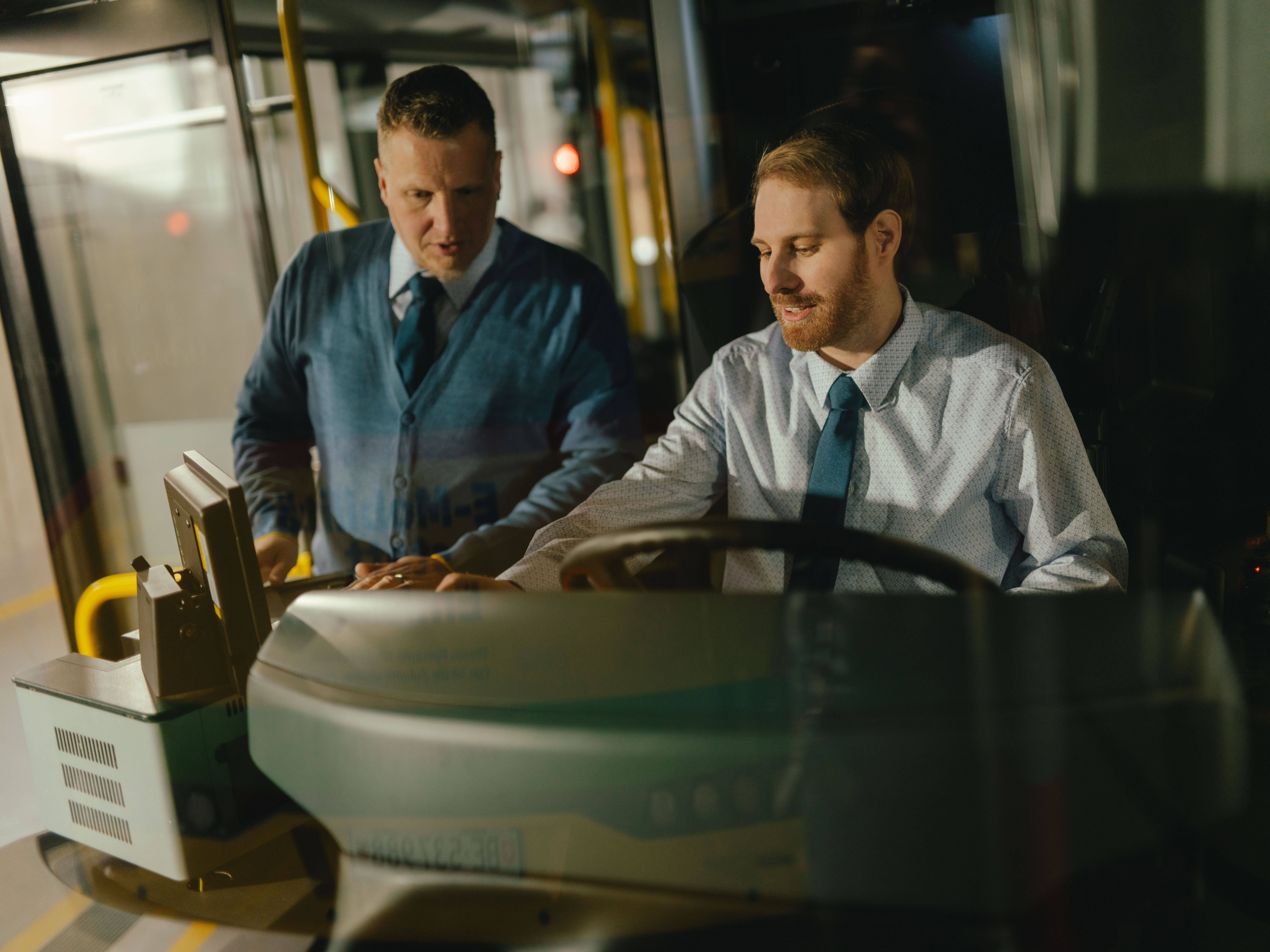 Two people are positioned in the driver’s area of a vehicle. One person is seated at the wheel while the other stands beside them. Both are wearing formal clothing. In the foreground, the steering wheel, control elements, and part of the dashboard are visible. The setting appears to be the interior of a bus.