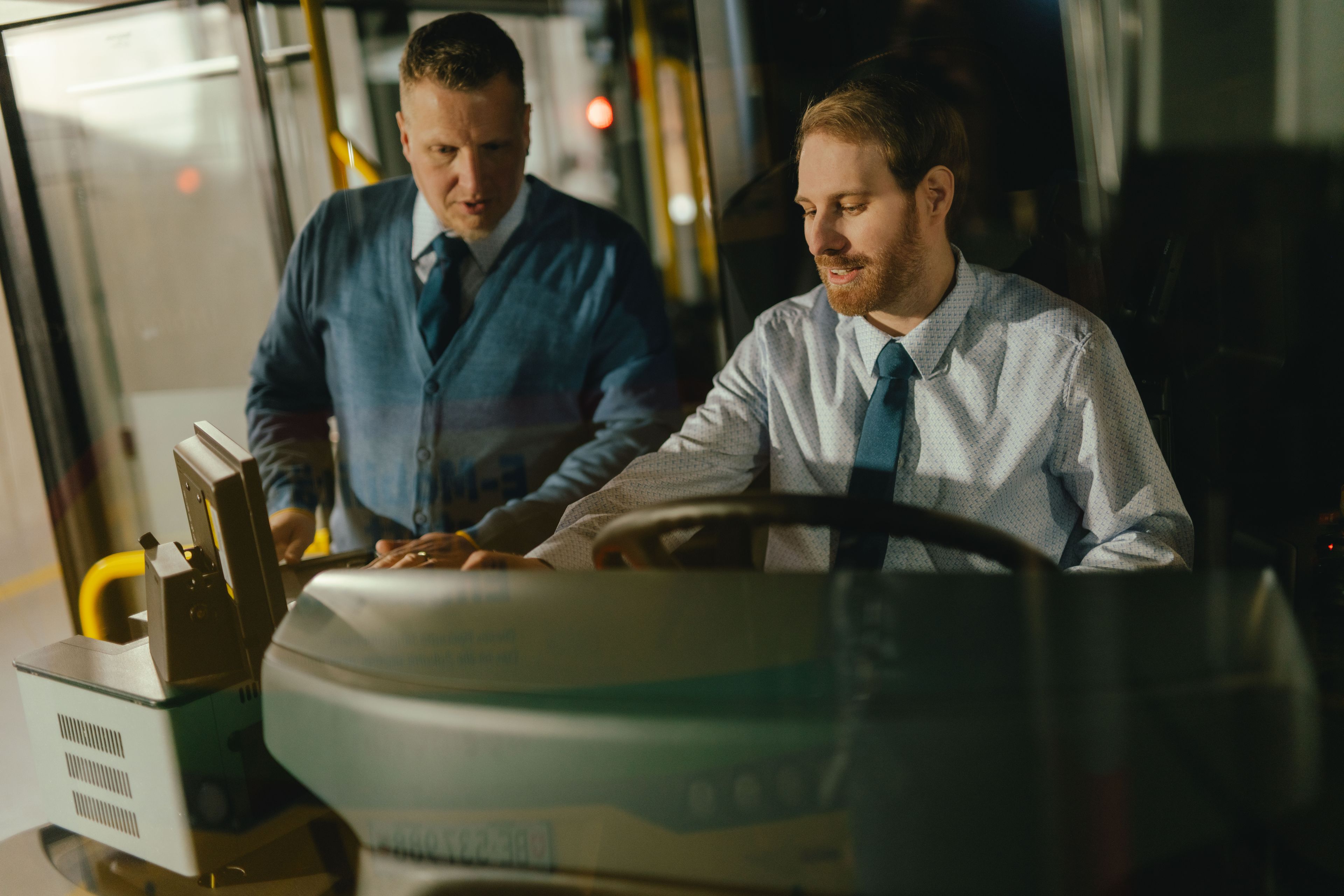 Two people are positioned in the driver’s area of a vehicle. One person is seated at the wheel while the other stands beside them. Both are wearing formal clothing. In the foreground, the steering wheel, control elements, and part of the dashboard are visible. The setting appears to be the interior of a bus.