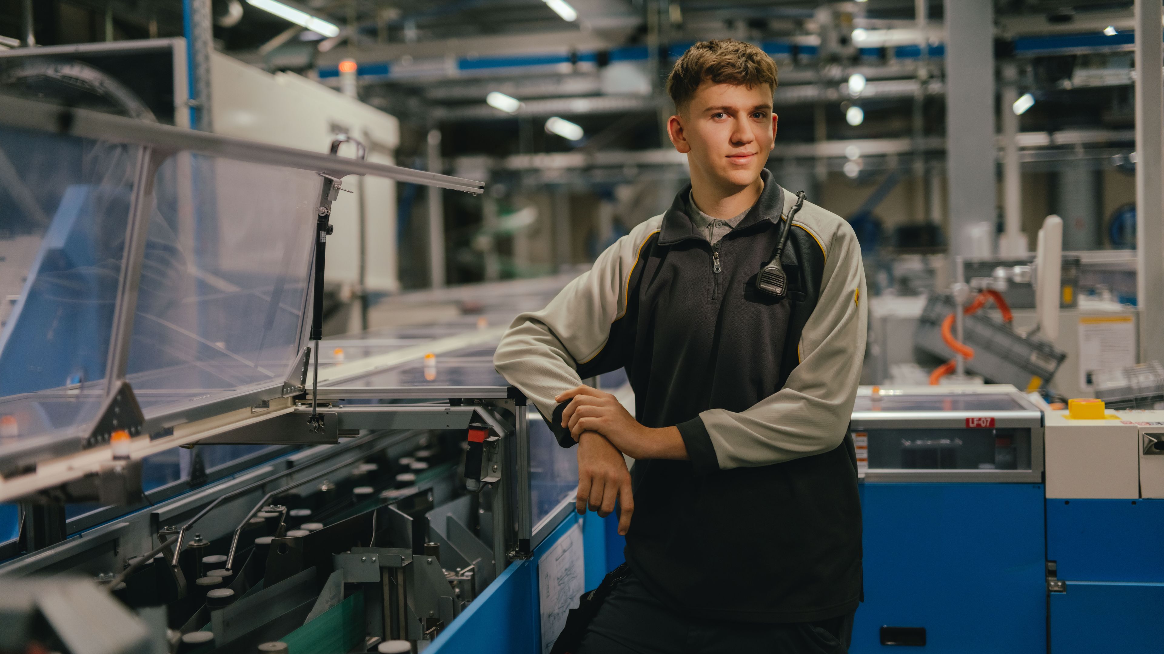 Eine Person steht in einer industriellen Halle zwischen grossen blauen Maschinen und technischen Anlagen. Die Person trägt dunkelgraue Arbeitskleidung mit hellen Ärmeln und lehnt sich mit einem Arm an eine Maschine. Im Hintergrund sind Förderanlagen, Kabel und Metallstrukturen sichtbar.