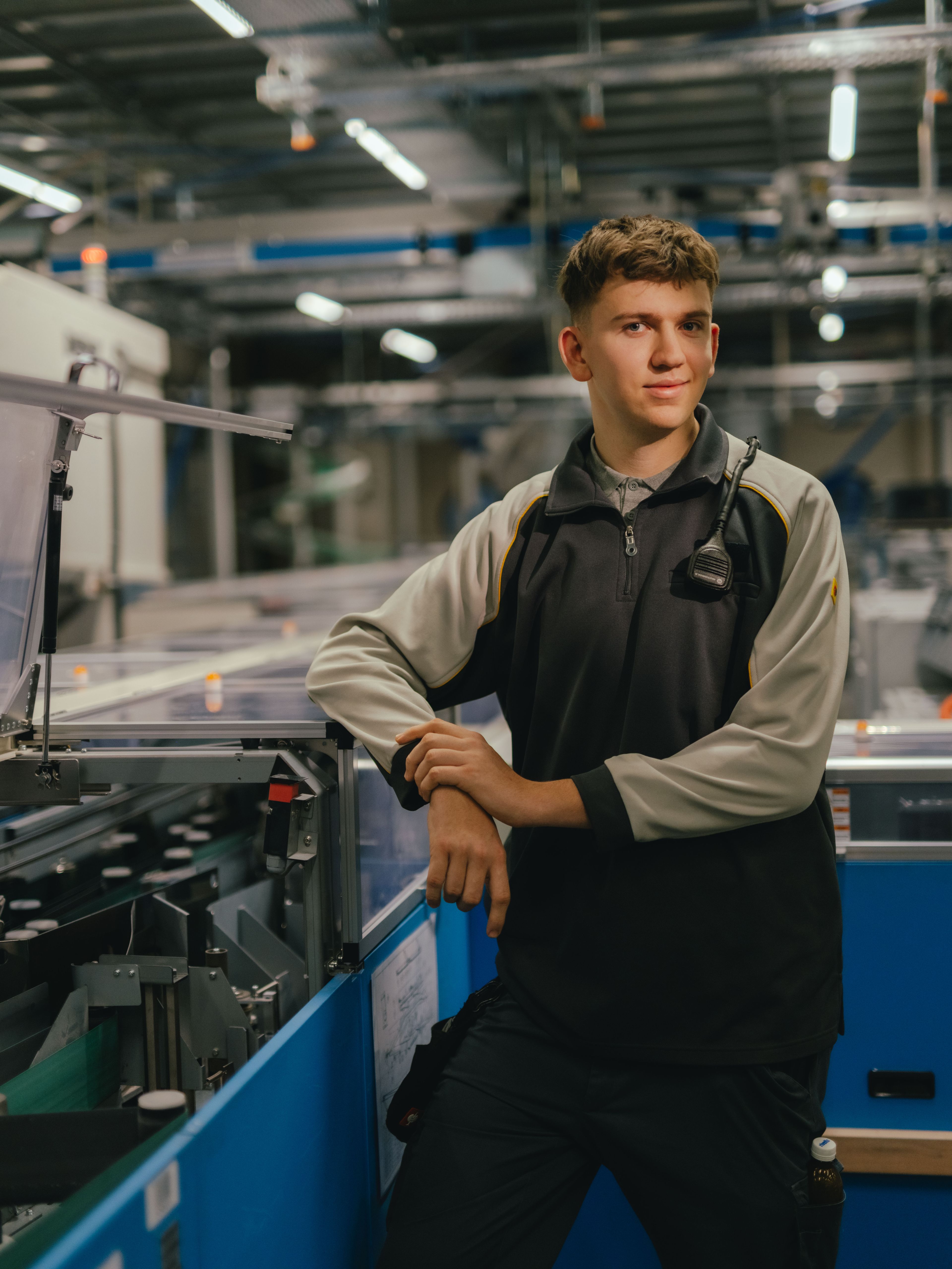 Une personne se tient dans une halle industrielle et s’appuie avec ses deux bras sur une machine équipée de capots transparents. La personne porte une veste de travail bicolore. En arrière-plan, d’autres machines, structures métalliques et éléments d’éclairage de l’environnement de production sont visibles.