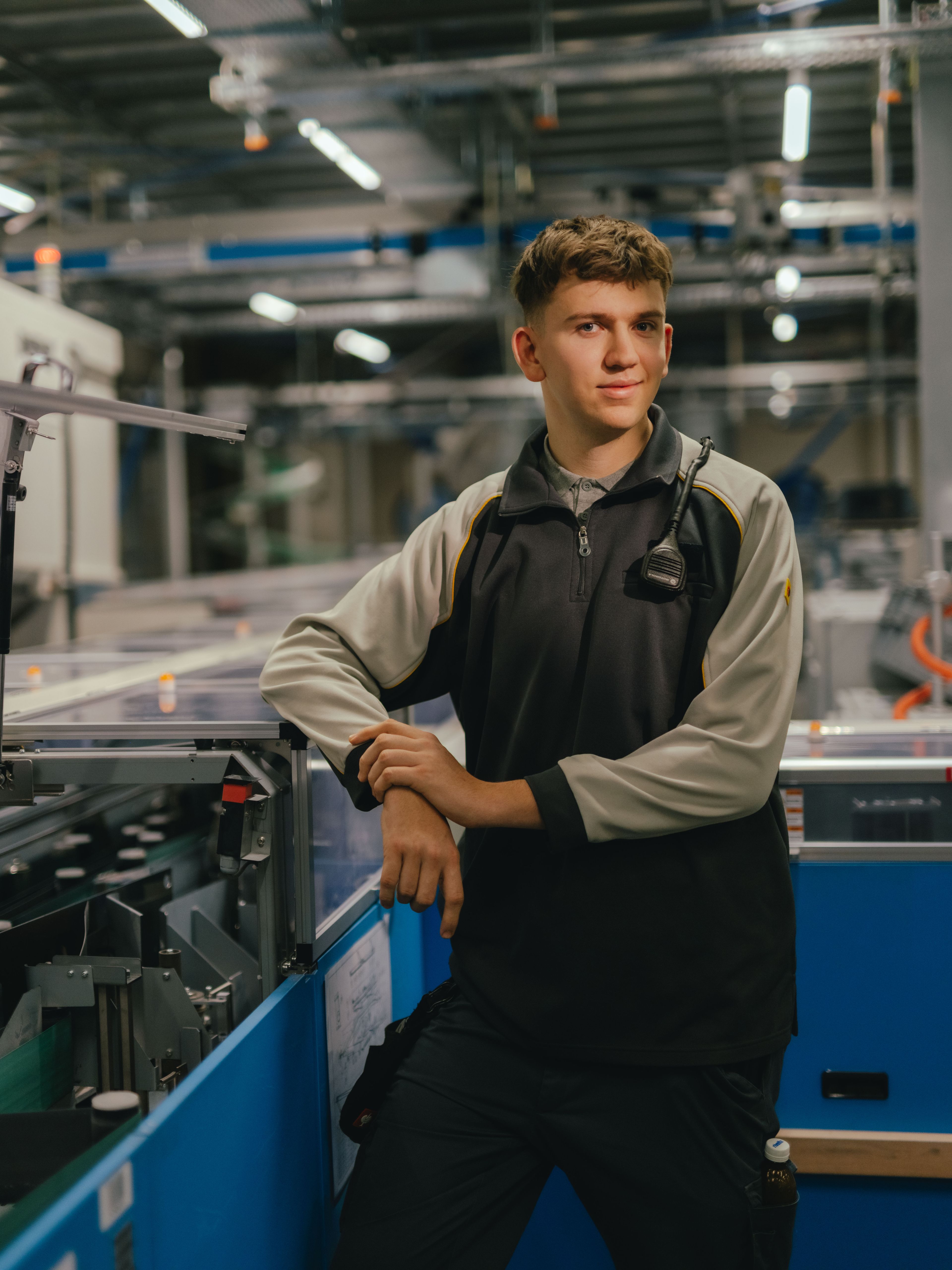 A person is standing in an industrial hall and leaning with both arms on a machine with transparent covers. They are wearing a two-coloured work jacket. Other machines, metal structures and lighting elements from the production environment can be seen in the background.