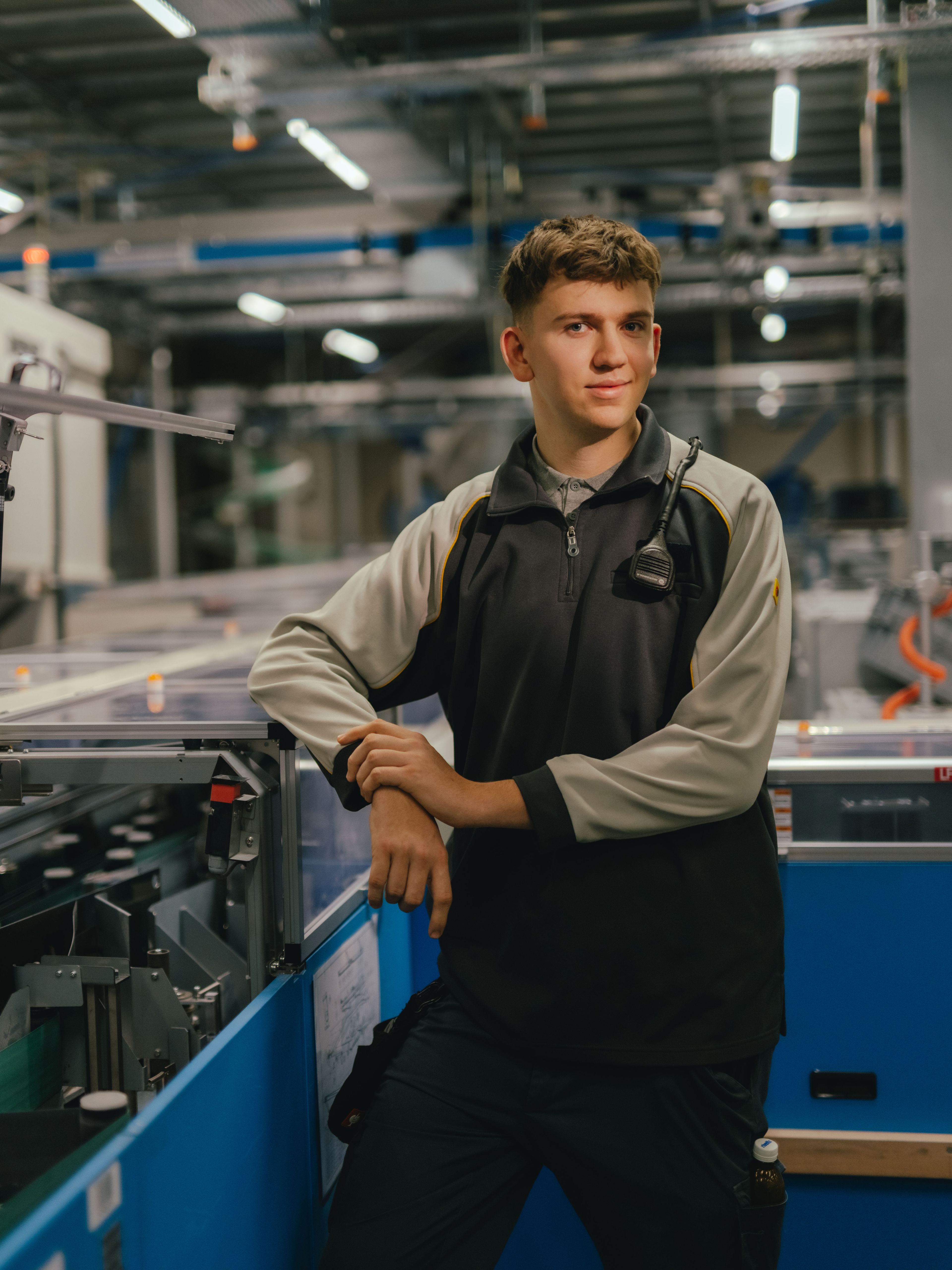 Eine Person steht in einer industriellen Halle und lehnt sich mit beiden Armen auf eine Maschine mit transparenten Abdeckungen. Die Person trägt eine zweifarbige Arbeitsjacke. Im Hintergrund sind weitere Maschinen, Metallstrukturen und Beleuchtungselemente der Produktionsumgebung sichtbar.