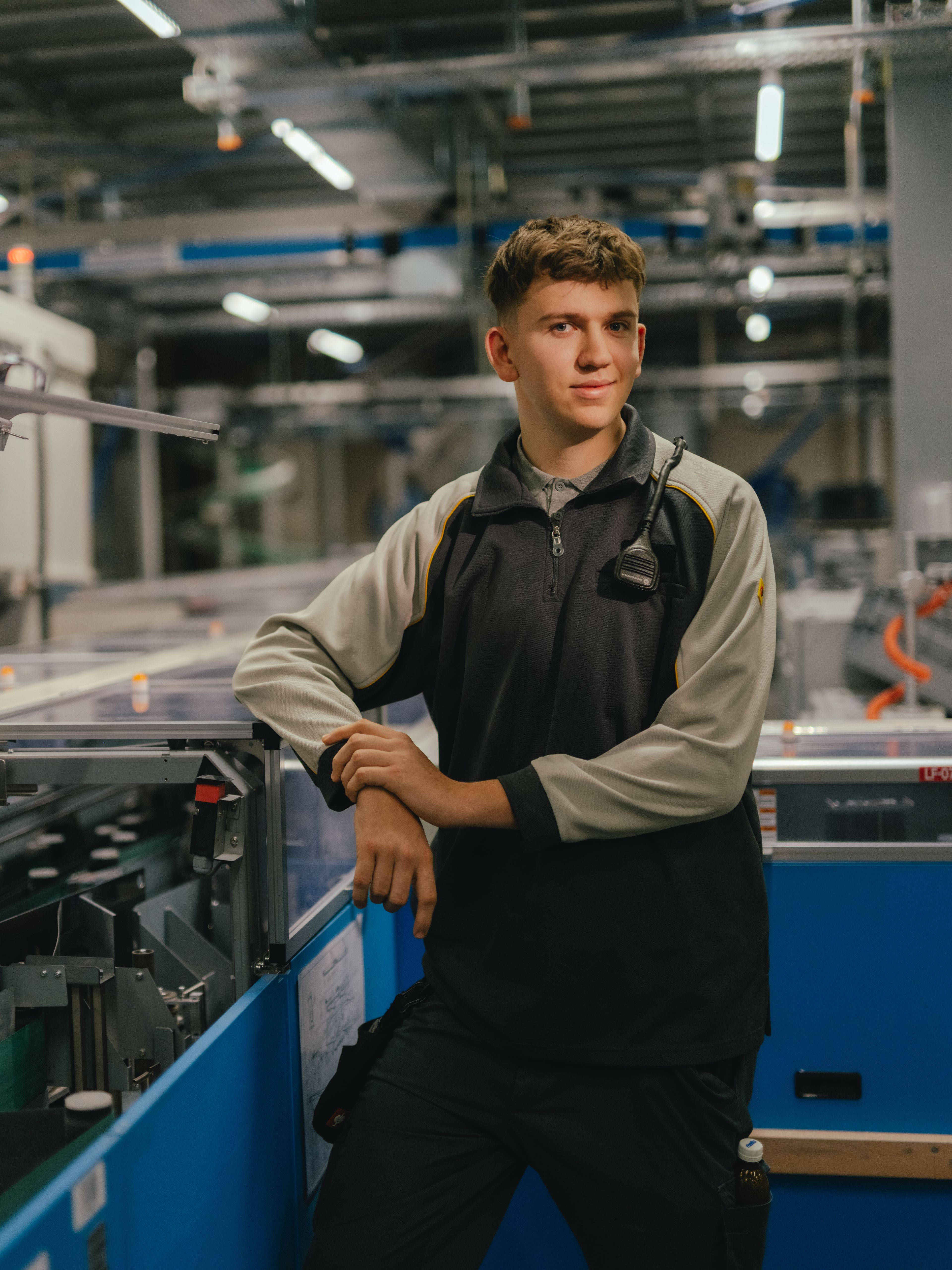 Una persona è in piedi in un padiglione industriale e si appoggia con entrambe le braccia su una macchina con coperture trasparenti. La persona indossa una giacca da lavoro bicolore. Sullo sfondo sono visibili altre macchine, strutture metalliche ed elementi di illuminazione dell’ambiente di produzione.