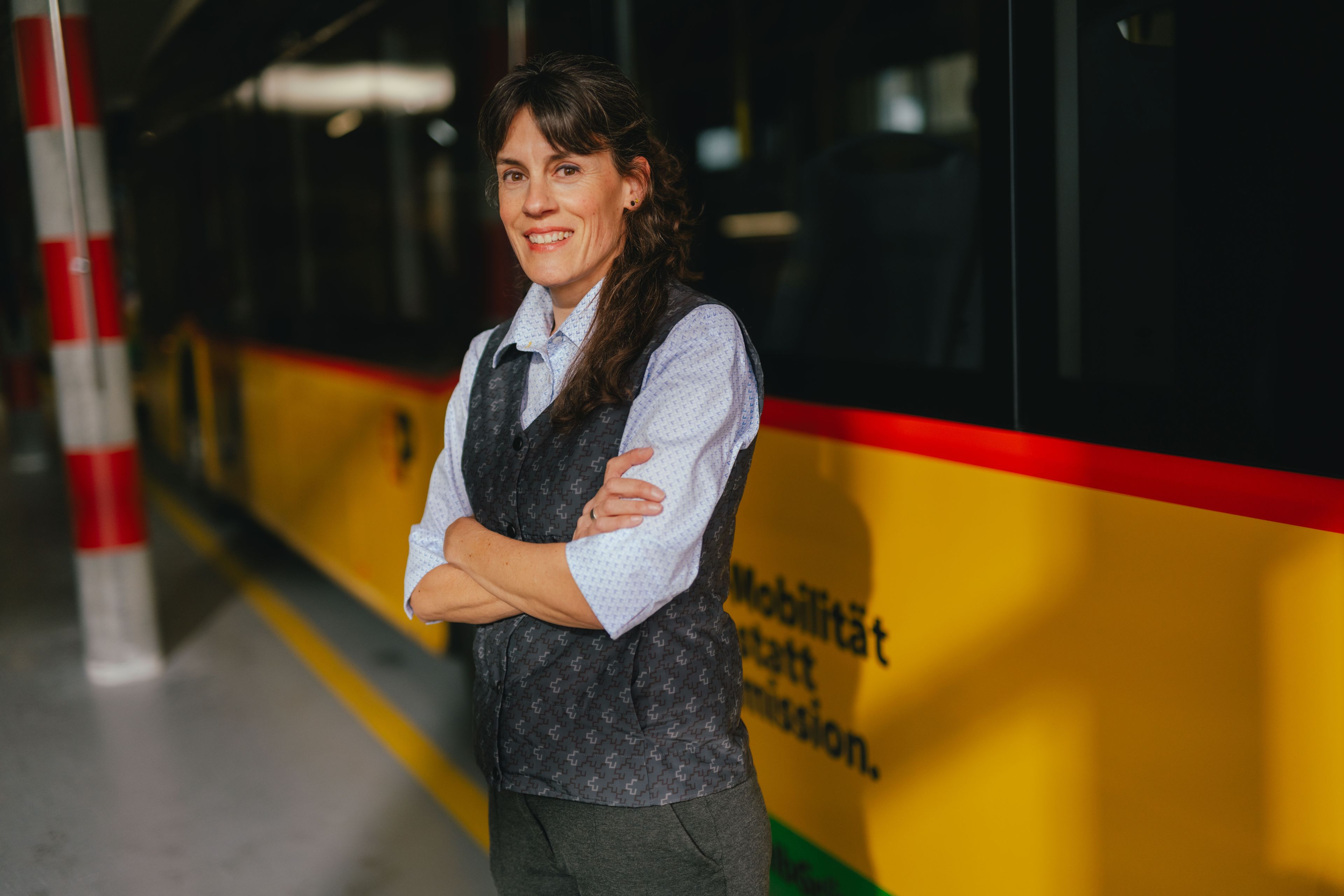 Une personne se tient les bras croisés dans un hall, à côté d’un véhicule jaune et noir. La personne porte une chemise claire et un gilet foncé. L’environnement ressemble à un dépôt ou une zone de stationnement, avec des colonnes rayées et un sol réfléchissant.