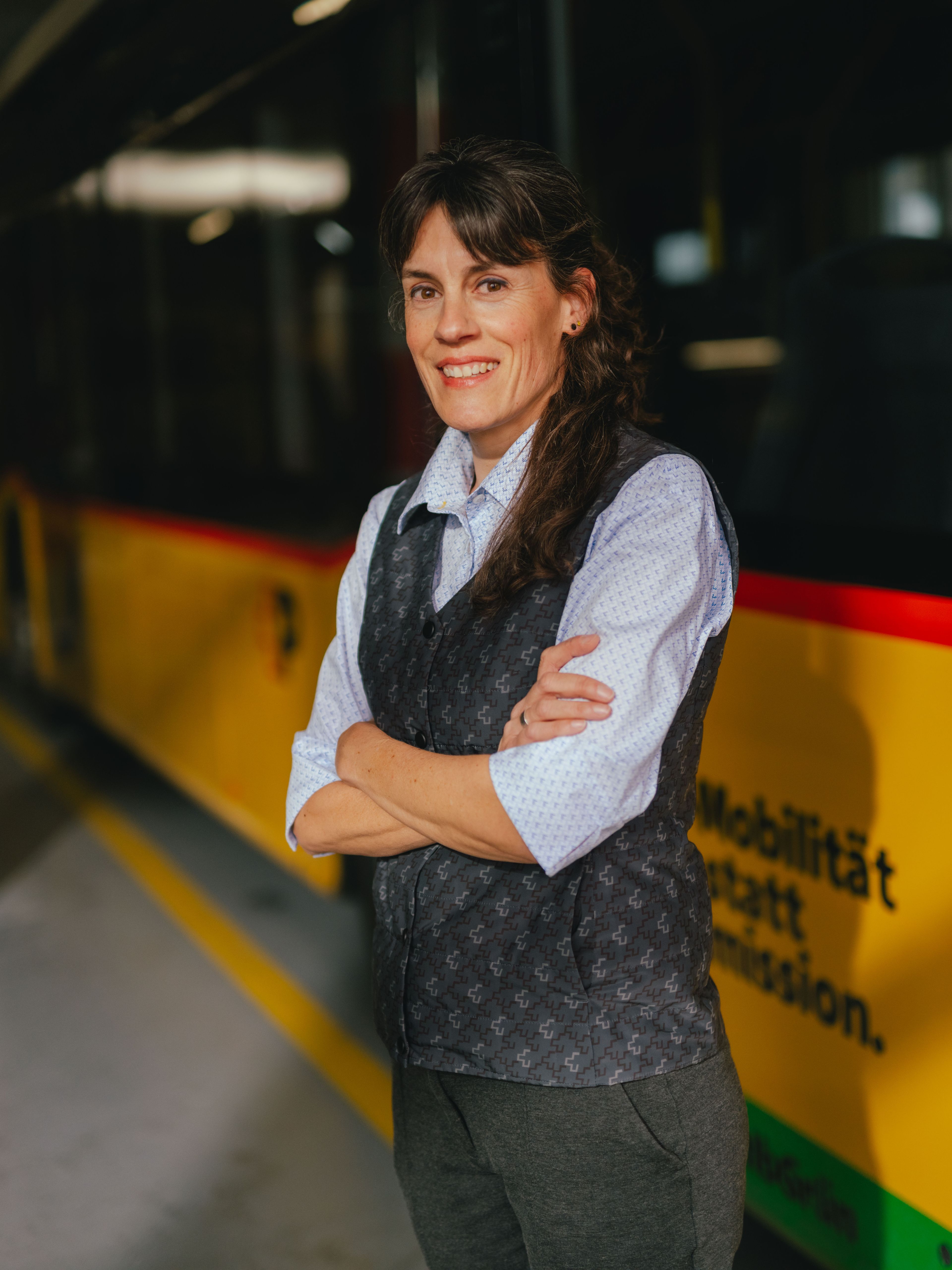 A person stands with arms crossed in a hall next to a yellow and black vehicle. The person is wearing a light shirt and a dark vest. The setting appears to be a depot or storage area, featuring striped pillars and a reflective floor.