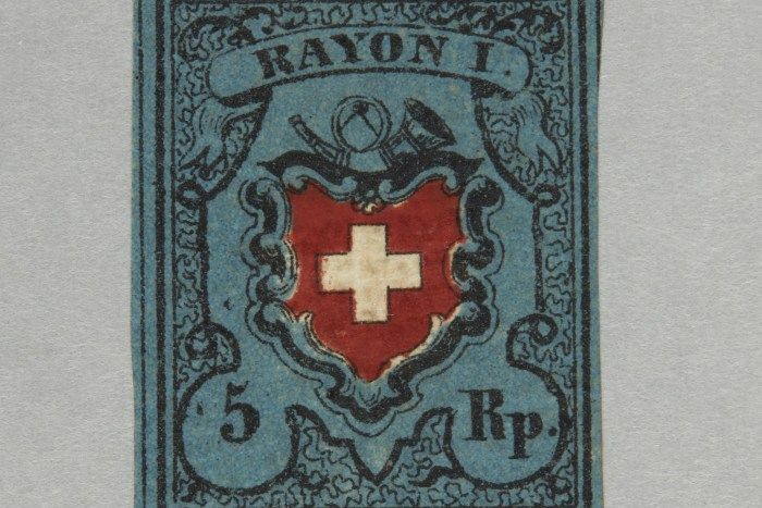 Historische Schweizer Briefmarke mit der Aufschrift ‚Rayon I‘. In der Mitte befindet sich ein rotes Wappenschild mit weissem Kreuz, umgeben von einer blauen dekorativen Umrandung. Oben sind Posthorn und Trompete abgebildet. Unten steht der Wert ‚5 Rp.‘.