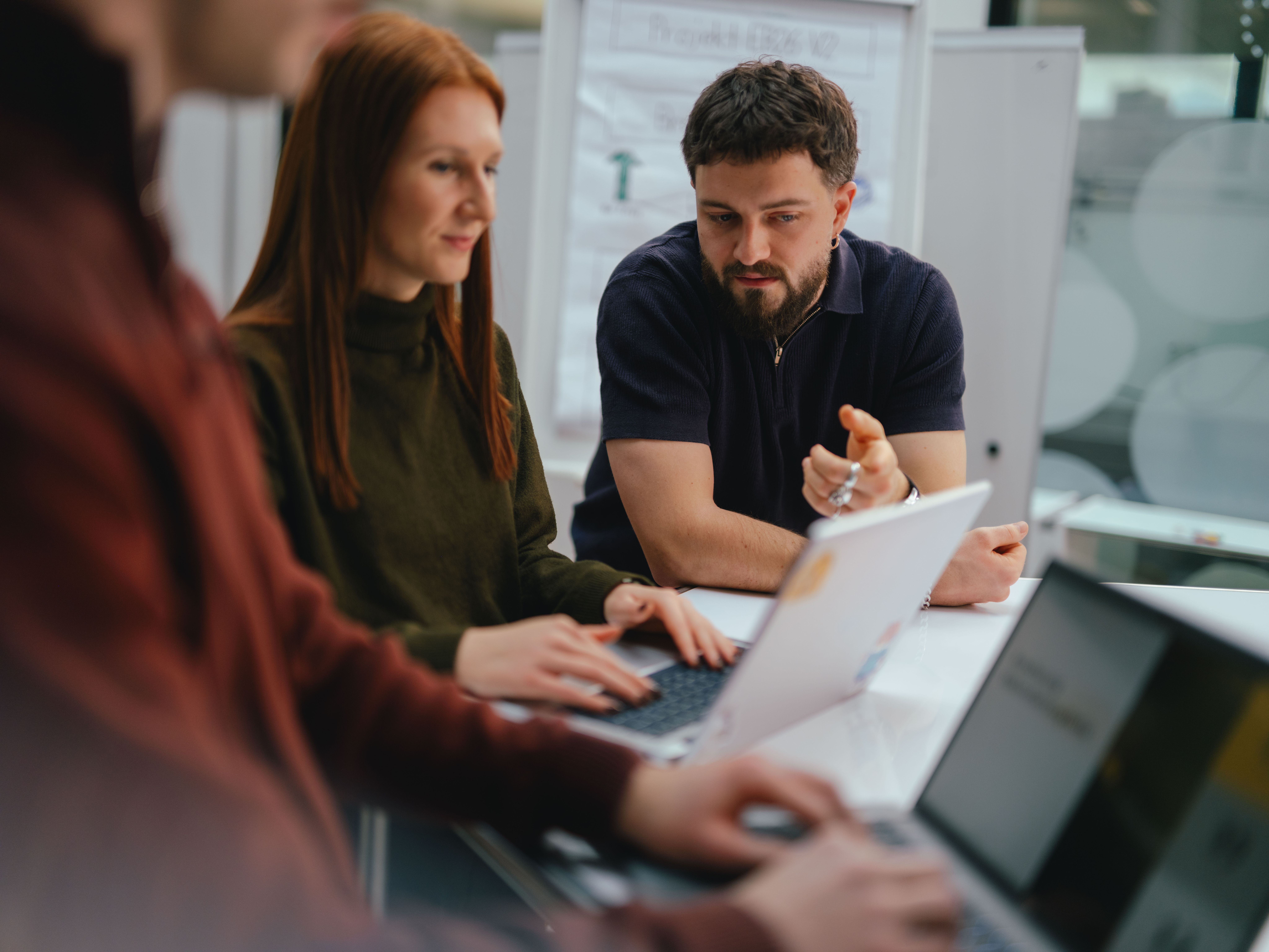 Mehrere Personen sitzen oder stehen an einem Tisch und arbeiten gemeinsam an Laptops. Eine Person im Vordergrund tippt auf einem Laptop, während zwei weitere Personen dahinter sitzen; eine von ihnen weist mit der Hand auf etwas auf dem Tisch oder Bildschirm hin. Auf dem Tisch stehen mehrere geöffnete Laptops sowie ein Glas Wasser. Im Hintergrund sind Glastrennwände und moderne Büroeinrichtung sichtbar.