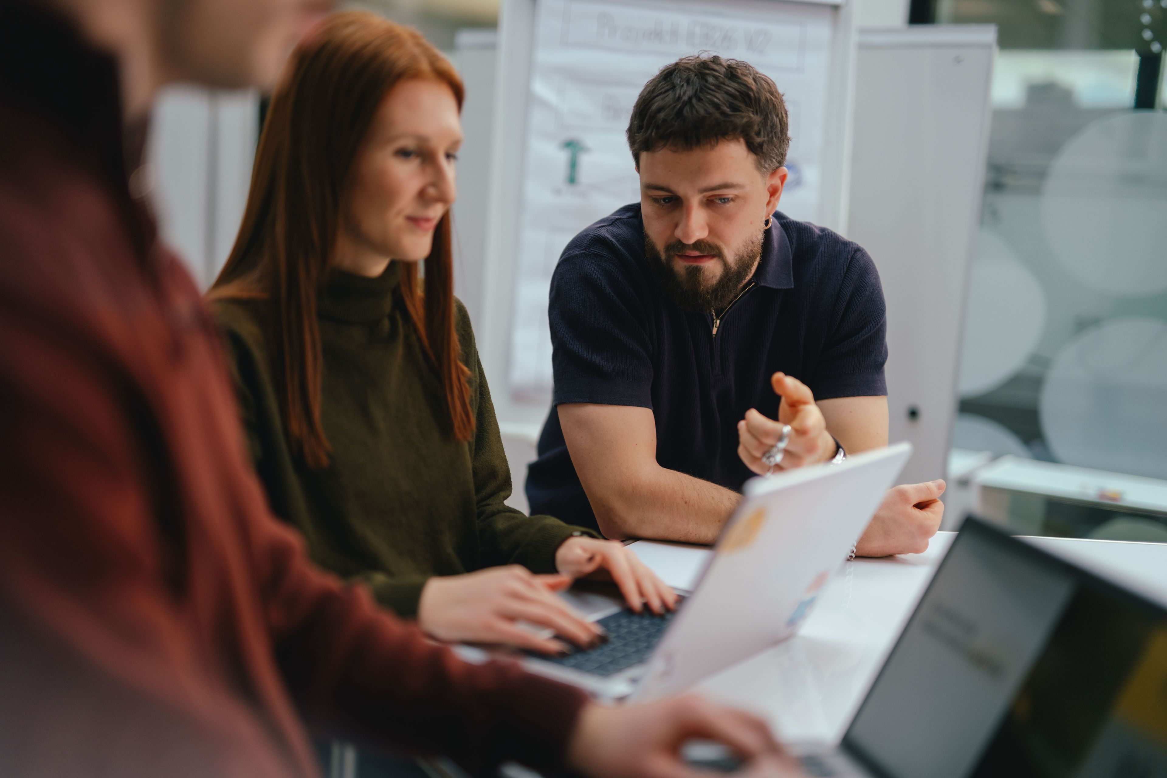 Mehrere Personen sitzen oder stehen an einem Tisch und arbeiten gemeinsam an Laptops. Eine Person im Vordergrund tippt auf einem Laptop, während zwei weitere Personen dahinter sitzen; eine von ihnen weist mit der Hand auf etwas auf dem Tisch oder Bildschirm hin. Auf dem Tisch stehen mehrere geöffnete Laptops sowie ein Glas Wasser. Im Hintergrund sind Glastrennwände und moderne Büroeinrichtung sichtbar.