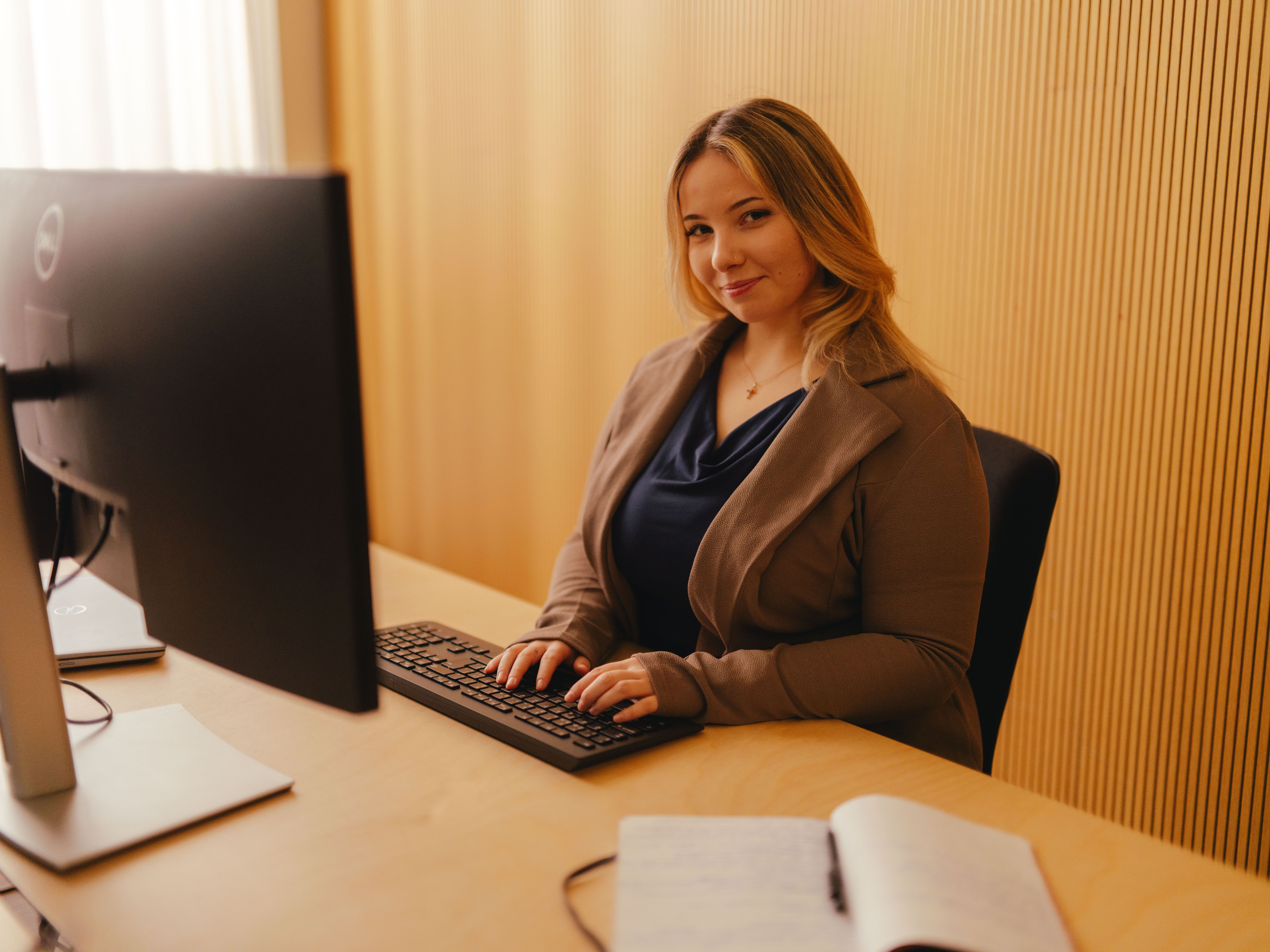 Eine Person sitzt an einem Schreibtisch und arbeitet an einem grossen Computerbildschirm. Die Person trägt eine dunkle Jacke über einem dunklen Oberteil und hat beide Hände auf der Tastatur. Auf dem Tisch liegt ein geöffnetes Notizbuch. Im Hintergrund befindet sich eine Wand aus senkrechten Holzpaneelen, und von links fällt Tageslicht in den Raum.