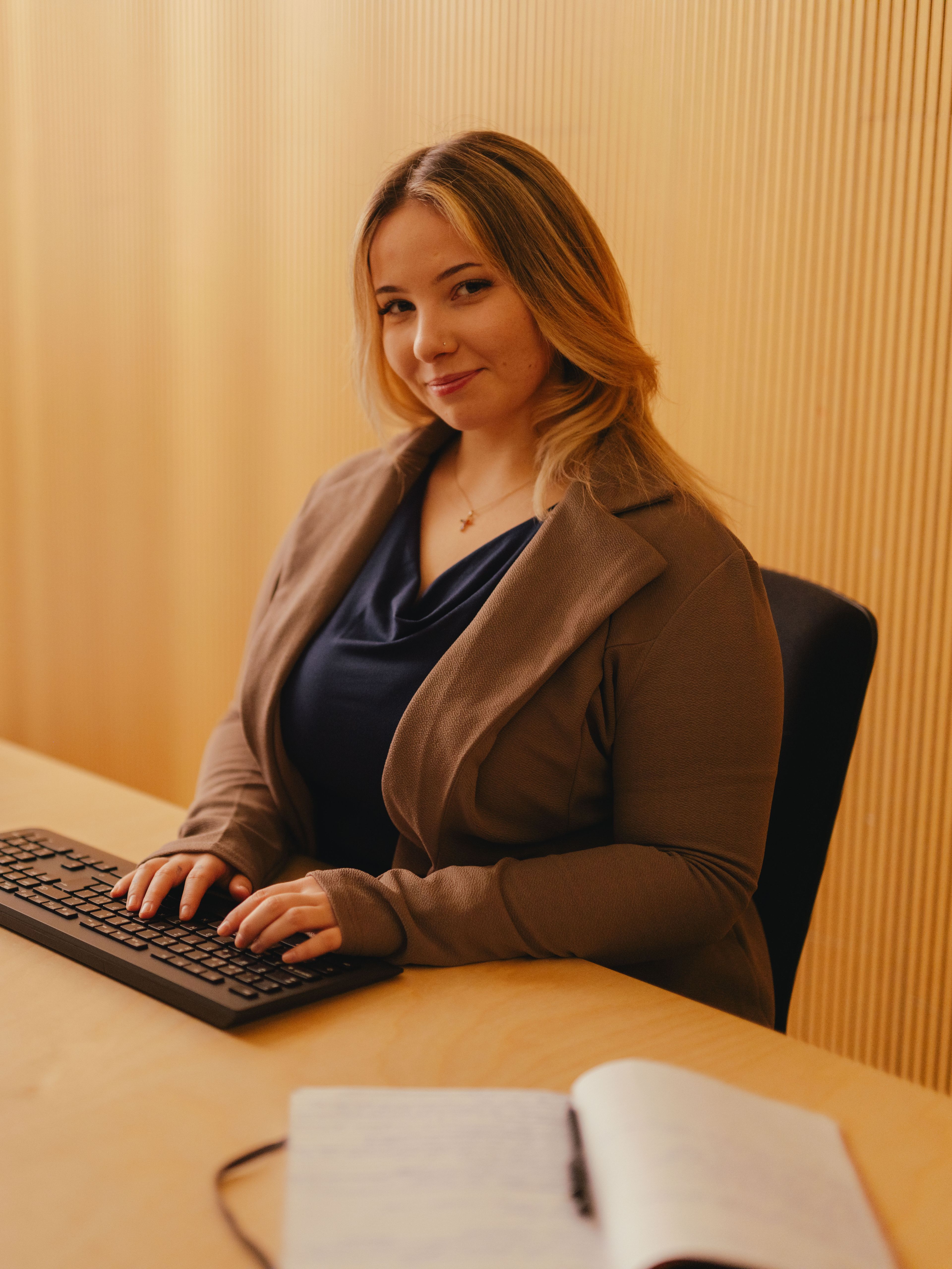 Eine Person sitzt an einem Schreibtisch und arbeitet an einem grossen Computerbildschirm. Die Person trägt eine dunkle Jacke über einem dunklen Oberteil und hat beide Hände auf der Tastatur. Auf dem Tisch liegt ein geöffnetes Notizbuch. Im Hintergrund befindet sich eine Wand aus senkrechten Holzpaneelen, und von links fällt Tageslicht in den Raum.