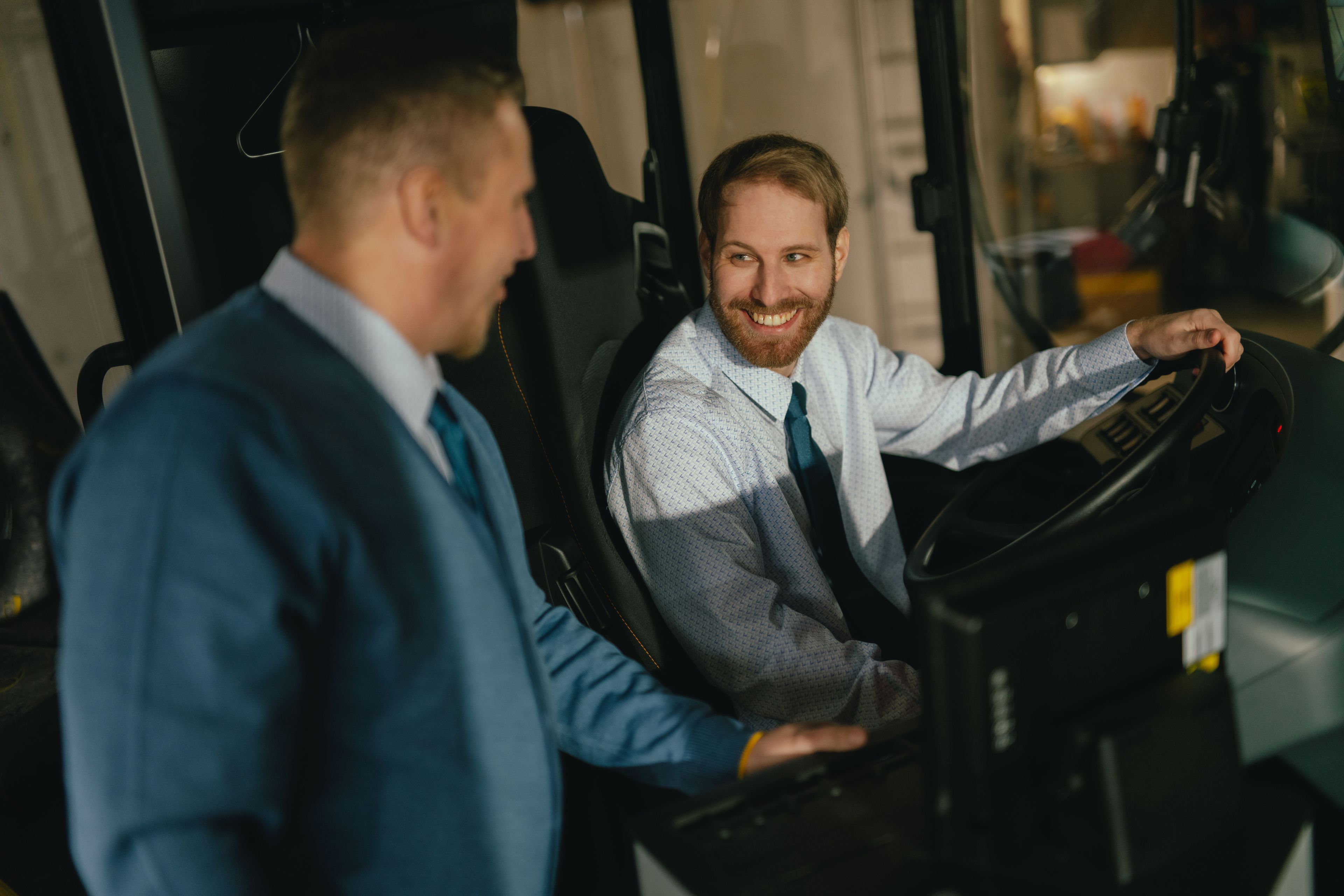 Two people are positioned in the driver’s area of a vehicle. One person is seated at the wheel, while the other stands beside them and leans slightly forward. Both are wearing formal clothing. The steering wheel, control elements, and part of the dashboard are visible. The setting appears to be the interior of a bus.