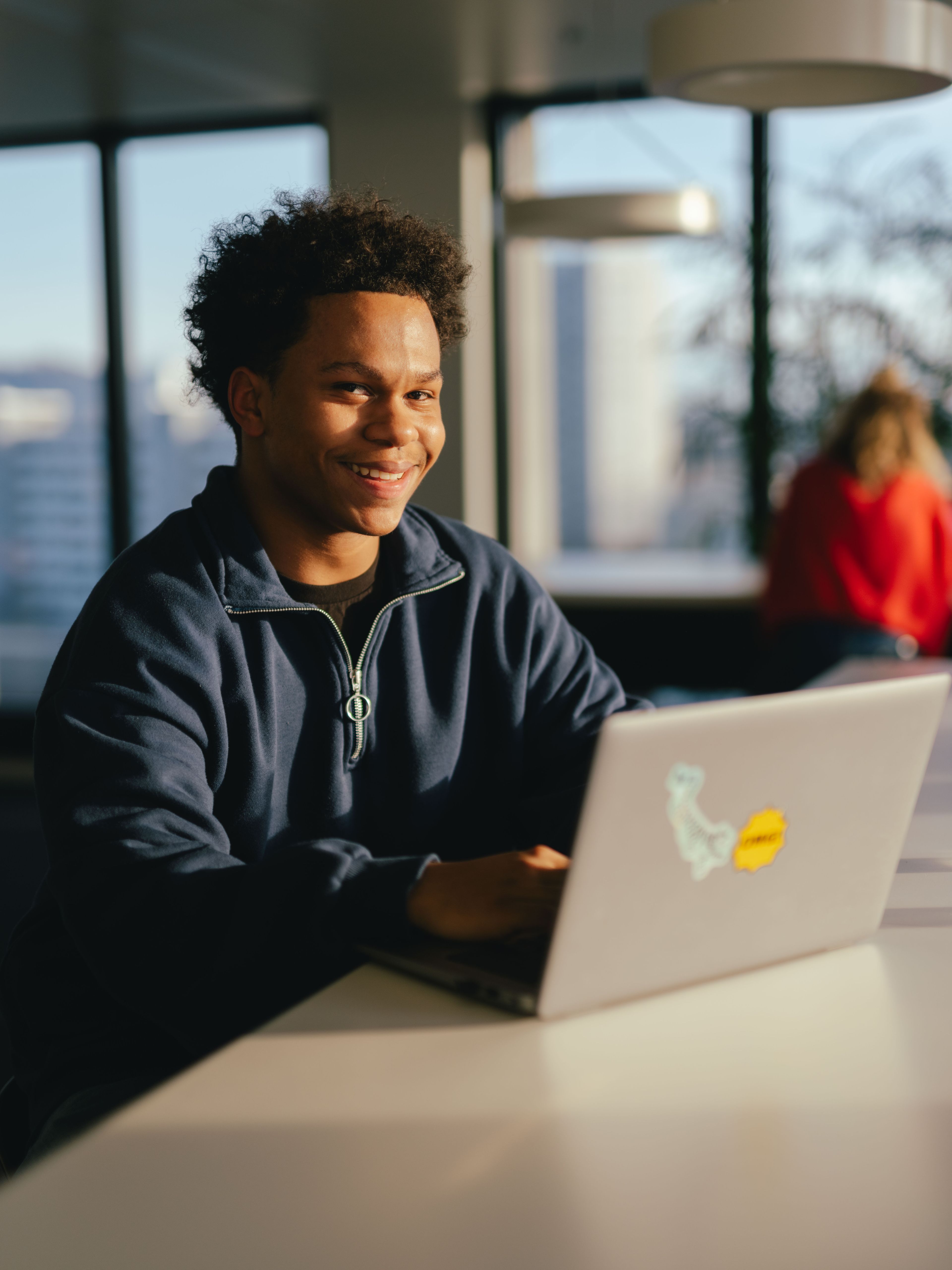 Eine Person sitzt an einem langen weissen Tisch und arbeitet an einem aufgeklappten Laptop, auf dessen Rückseite ein farbiges Aufklebermotiv zu sehen ist. Auf dem Tisch steht ausserdem eine durchsichtige Wasserflasche und ein leeres Glas. Der Raum ist hell, mit grossen Fenstern, durch die Tageslicht fällt. Im Hintergrund sind weitere Personen an einem Tisch sowie Büroelemente und Lampen sichtbar.