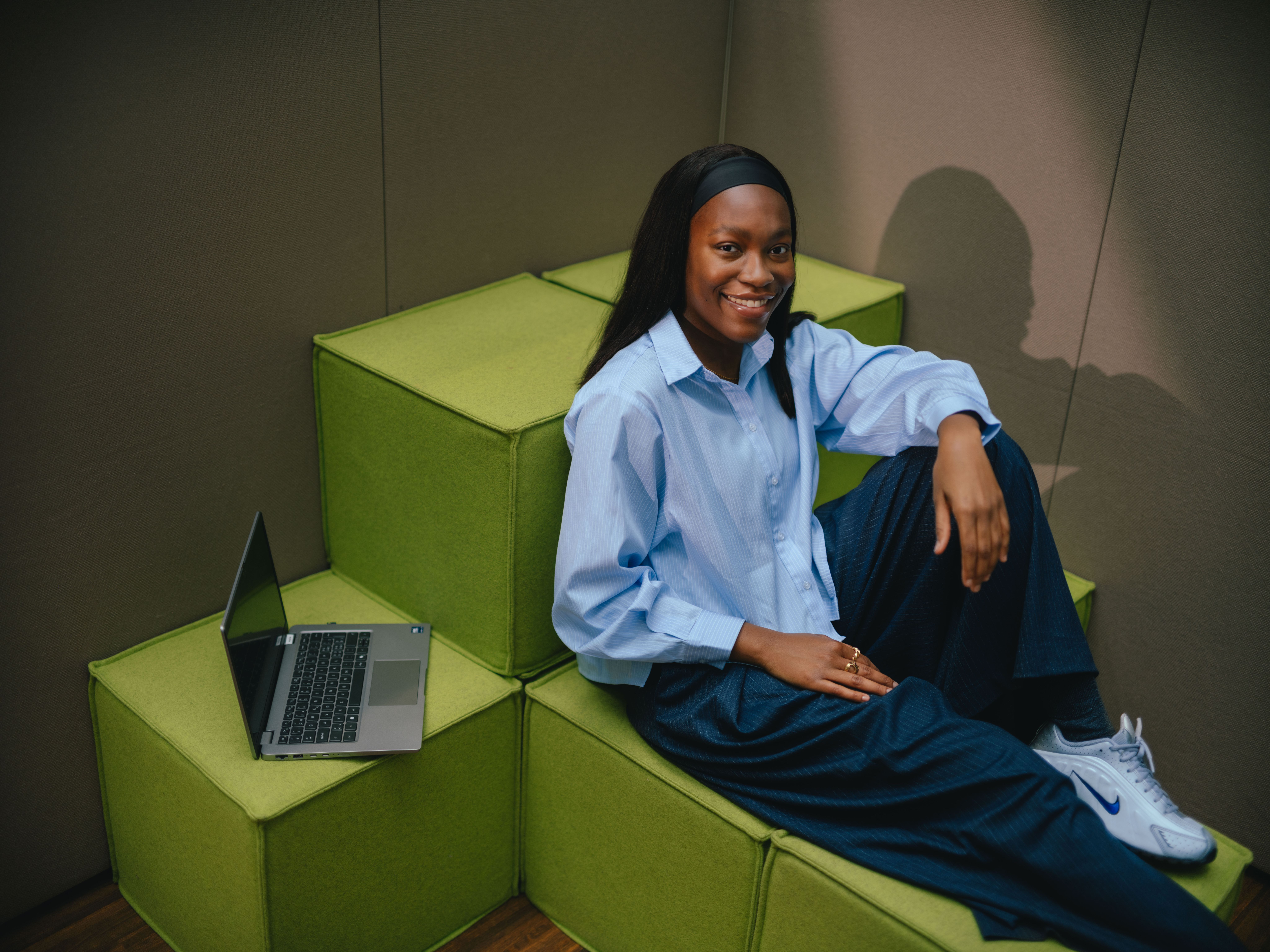 Eine Person sitzt an einem Tisch vor einem aufgeklappten Laptop. Die Person lehnt sich leicht zurück, während eine Hand auf dem Laptop liegt. Der Hintergrund besteht aus bodenlangen, beigefarbenen Vorhängen.