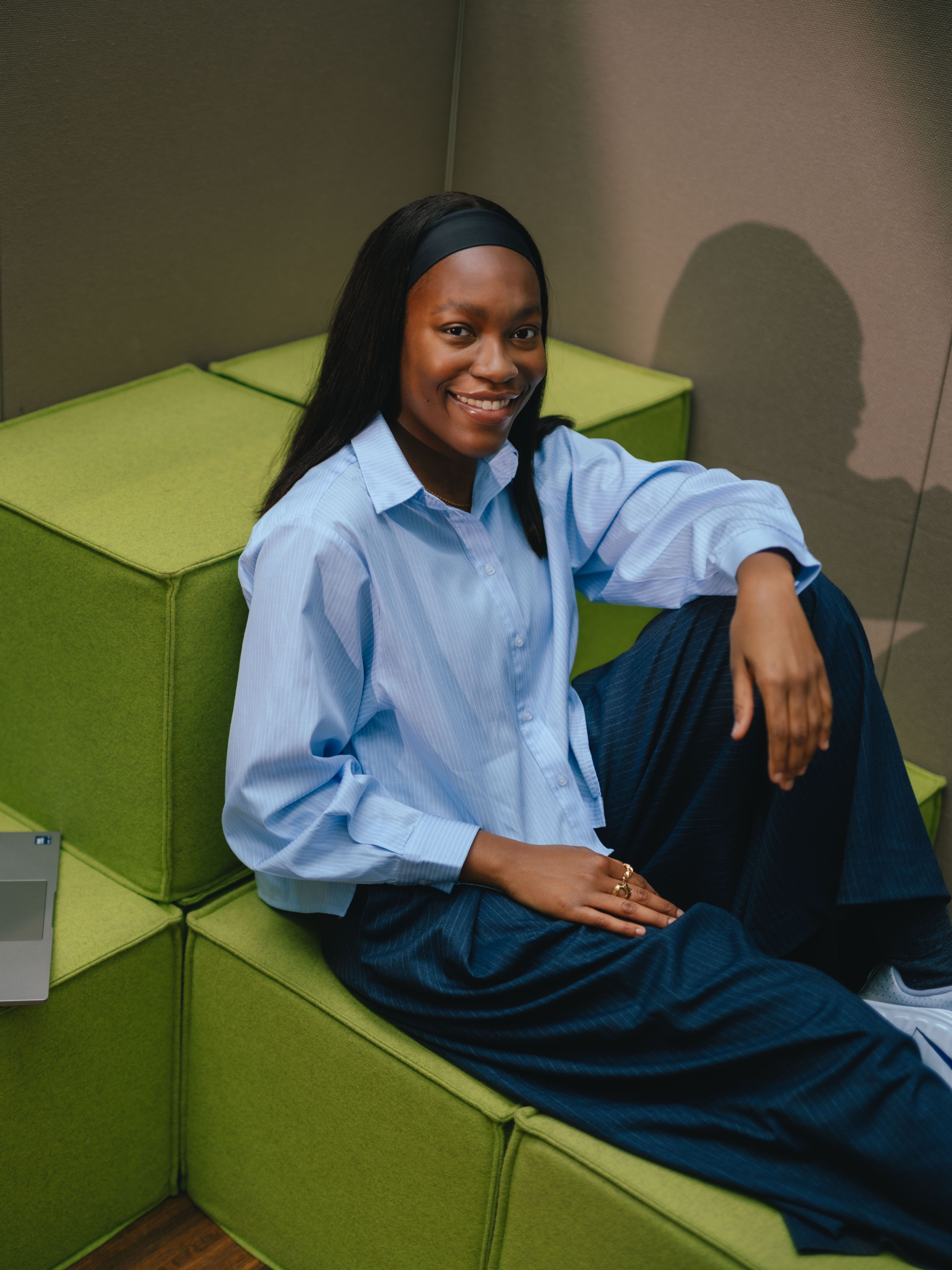 Una persona è seduta a un tavolo in una stanza con tende beige a tutta altezza. Davanti alla persona c’è un computer portatile aperto. La persona indossa una camicia azzurra ampia e si appoggia leggermente all’indietro, con un braccio poggiato sul tavolo.