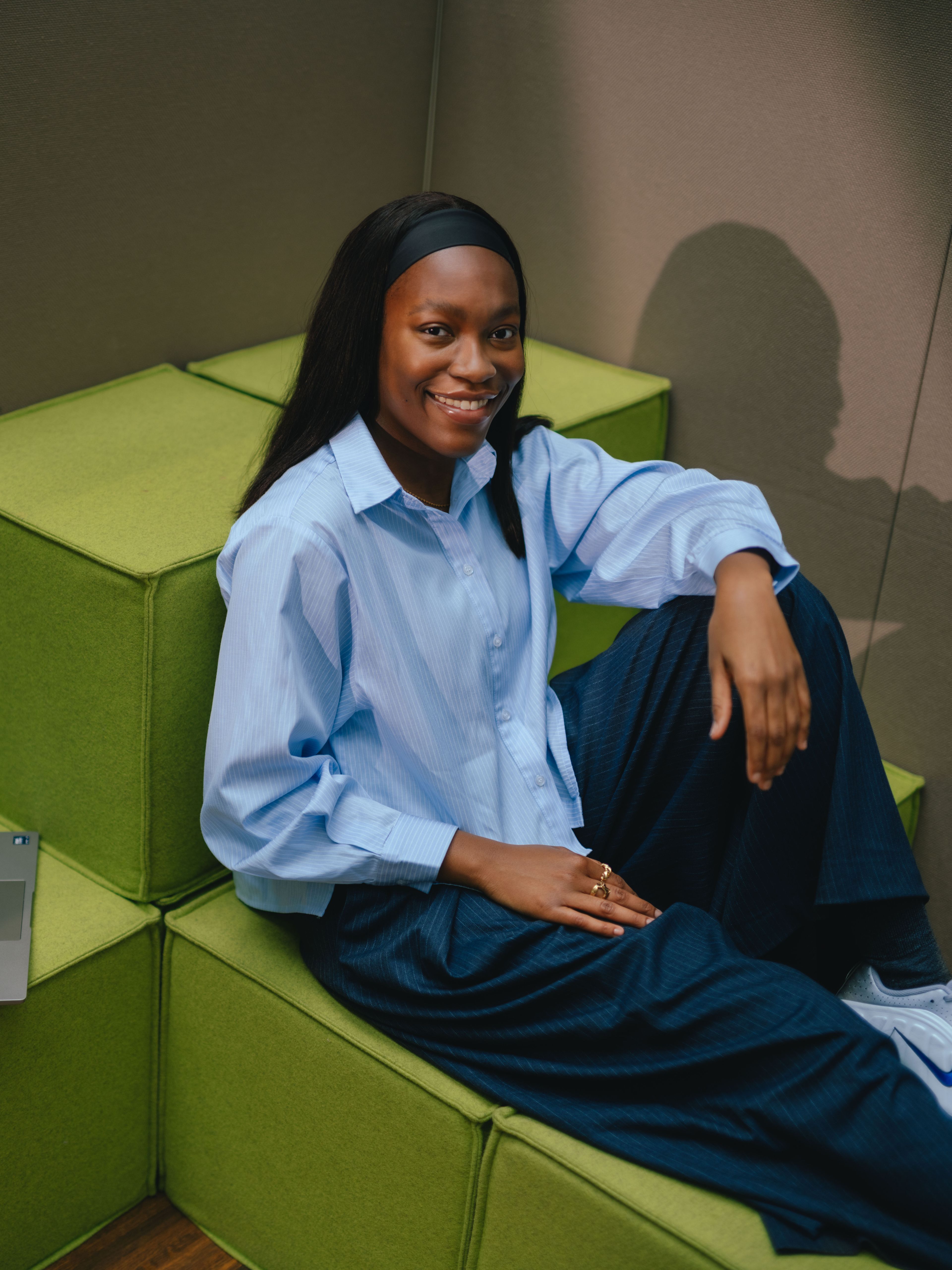 Eine Person sitzt an einem Tisch vor einem aufgeklappten Laptop. Die Person trägt ein hellblaues, locker geschnittenes Oberteil mit weiten Ärmeln. Im Hintergrund hängen bodenlange Vorhänge in einem warmen Beigeton, die den gesamten Raum gleichmässig ausleuchten. Die Haltung ist leicht zurückgelehnt, mit einem Arm auf dem Tisch abgestützt.