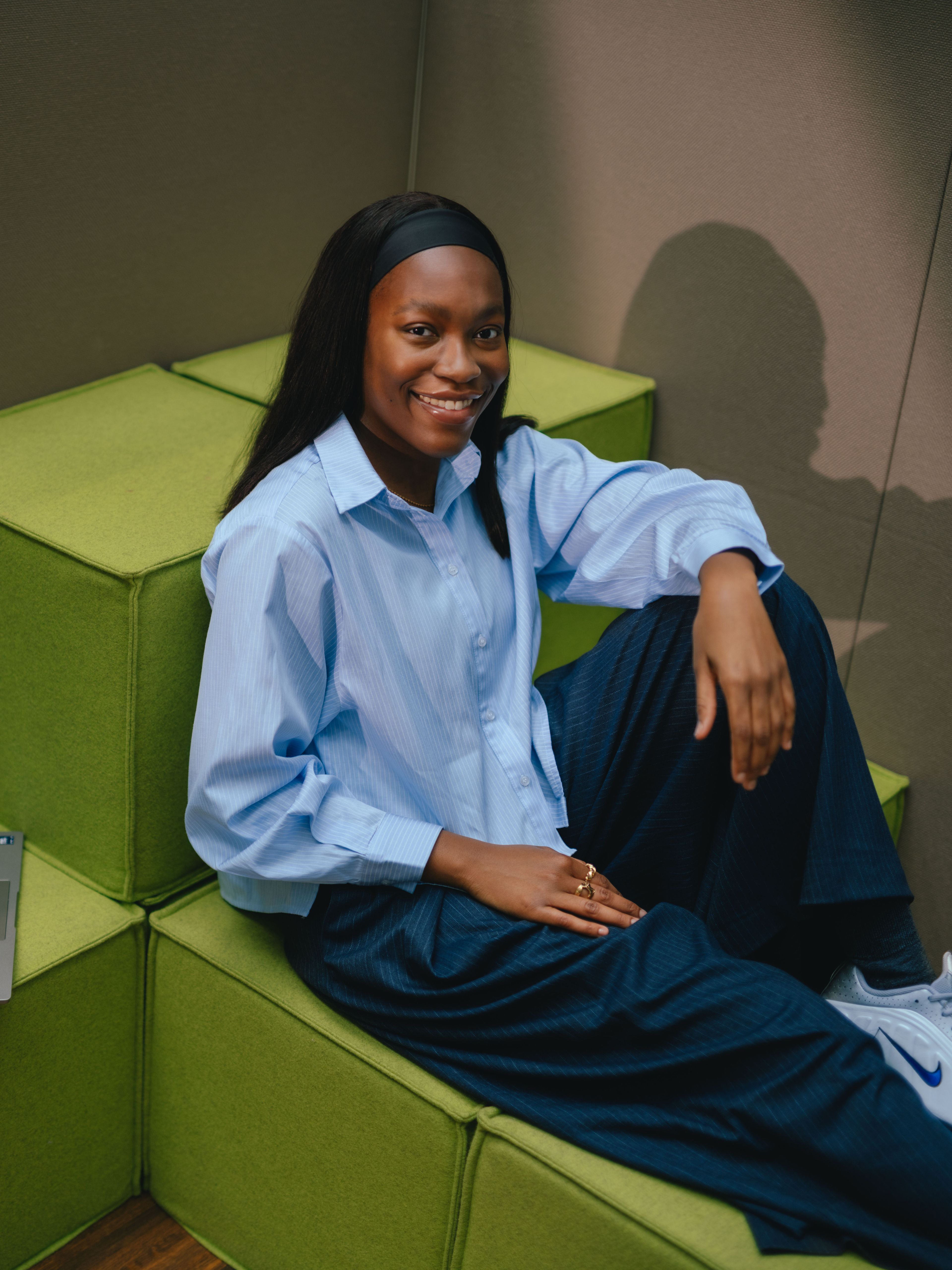 A person is seated at a table in a room with floor‑length beige curtains. An open laptop is placed on the table in front of the person. The person is wearing a loose light‑blue top and is leaning slightly back with one arm resting on the table.