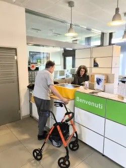 Une personne se tient à un comptoir d’accueil dans un bâtiment moderne. Devant elle, il y a un déambulateur orange. Derrière le comptoir, une autre personne travaille sur un ordinateur. Sur le comptoir se trouve un grand bac jaune. Le mot “Benvenuti” est écrit sur la façade du comptoir.