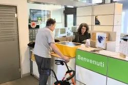 Une personne se tient à un comptoir d’accueil dans un bâtiment moderne. Devant elle, il y a un déambulateur orange. Derrière le comptoir, une autre personne travaille sur un ordinateur. Sur le comptoir se trouve un grand bac jaune. Le mot “Benvenuti” est écrit sur la façade du comptoir.