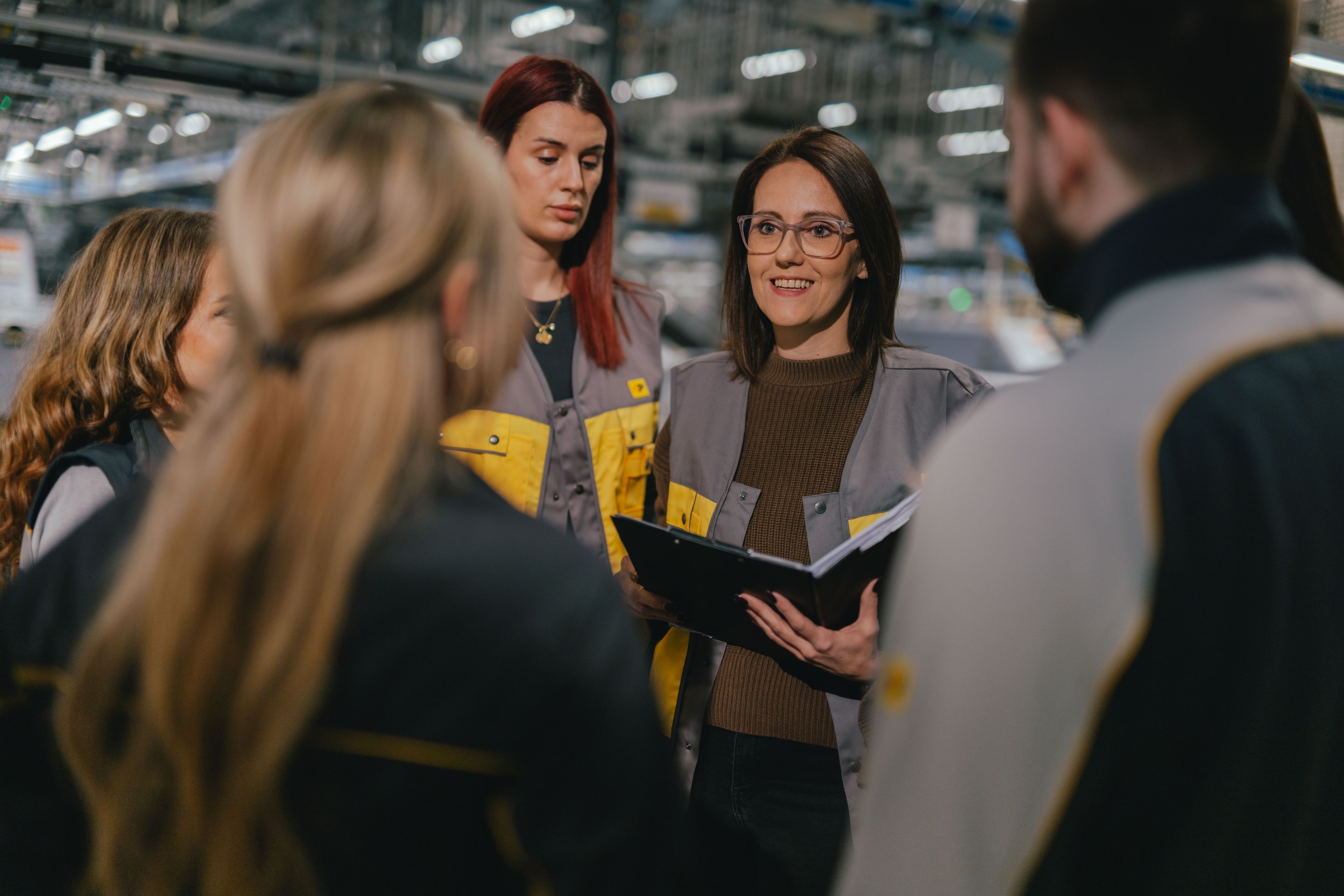 Un gruppo di persone è riunito in una sala di produzione o logistica. Diverse persone indossano abiti da lavoro grigi e gialli. Una persona tiene in mano un raccoglitore aperto mentre il gruppo è disposto in cerchio. Sullo sfondo si vedono macchinari, scaffalature e illuminazione intensa dal soffitto.