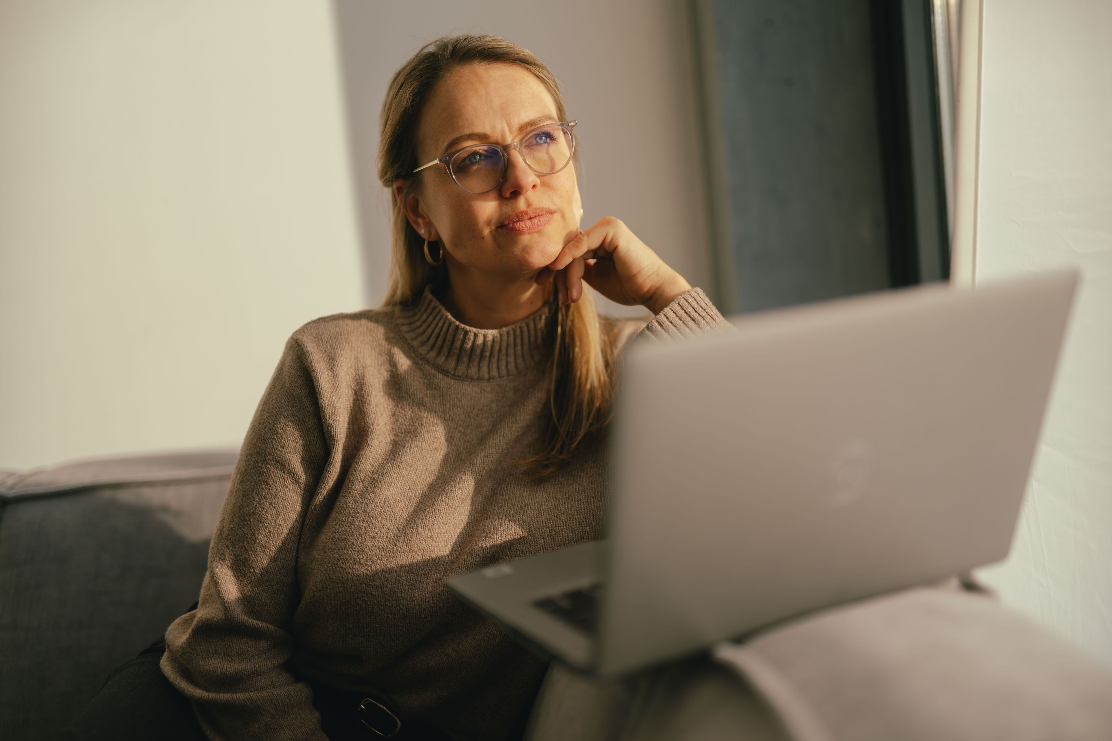 A person sits on a sofa near a window and works on an open laptop. The person is wearing a beige sweater, and warm sunlight illuminates the room.