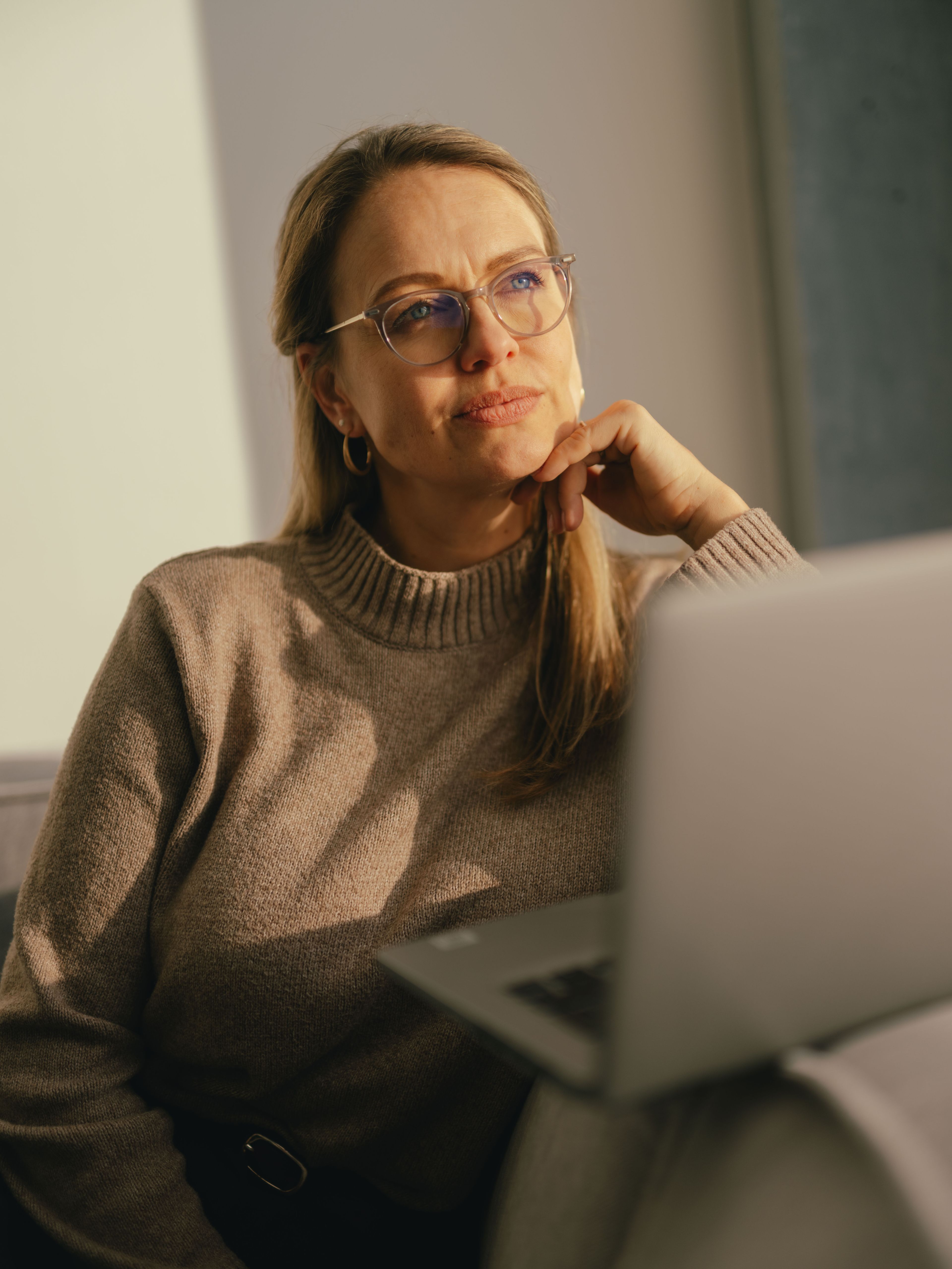 Une personne est assise sur un canapé près d’une fenêtre et travaille sur un ordinateur portable ouvert. La personne porte un pull beige, tandis qu’une lumière solaire chaude éclaire la pièce.