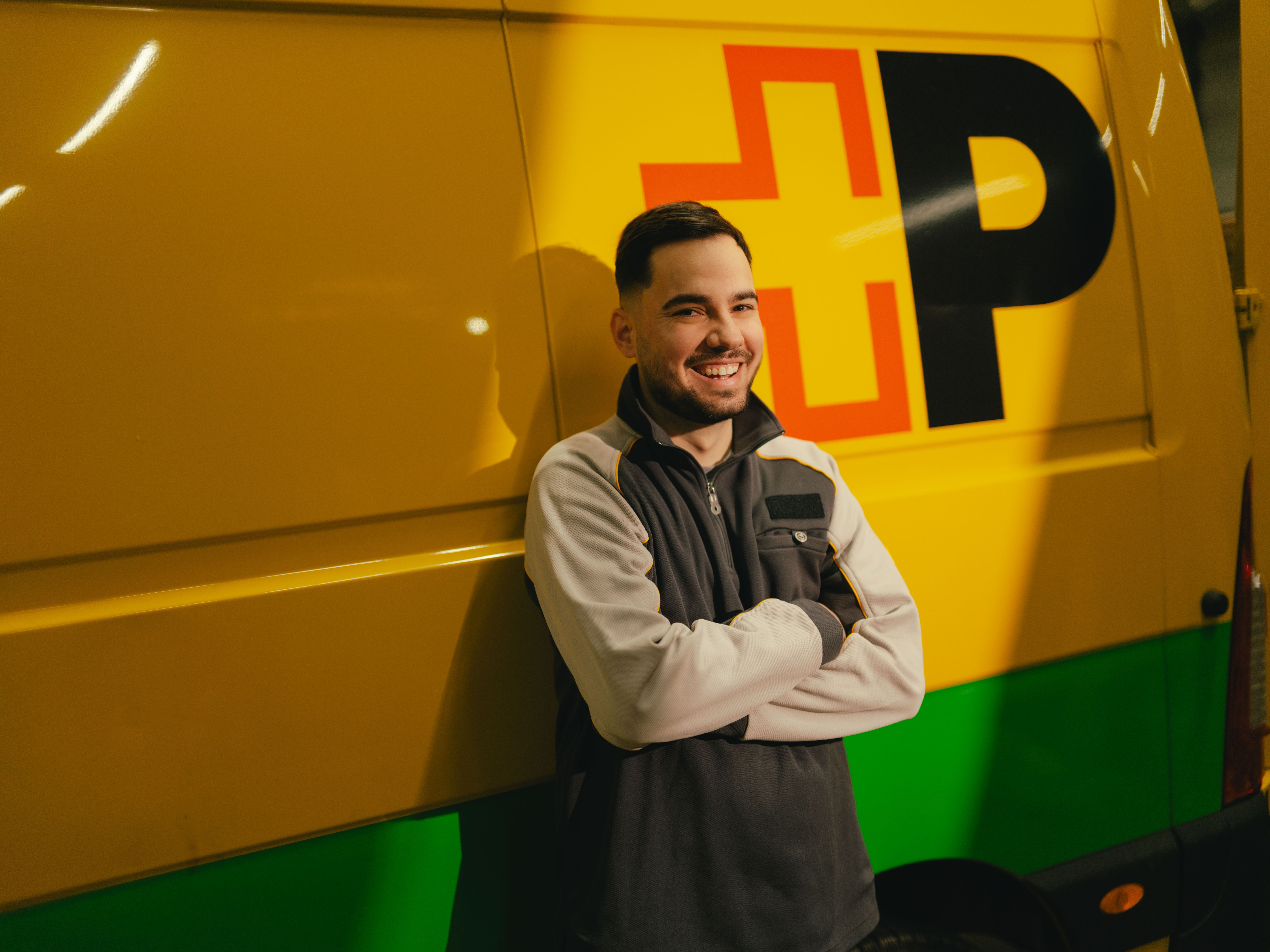 Eine Person steht mit verschränkten Armen vor einem gelb‑grünen Lieferfahrzeug mit grossen, schwarzen und roten Buchstaben auf der Fahrzeugseite.