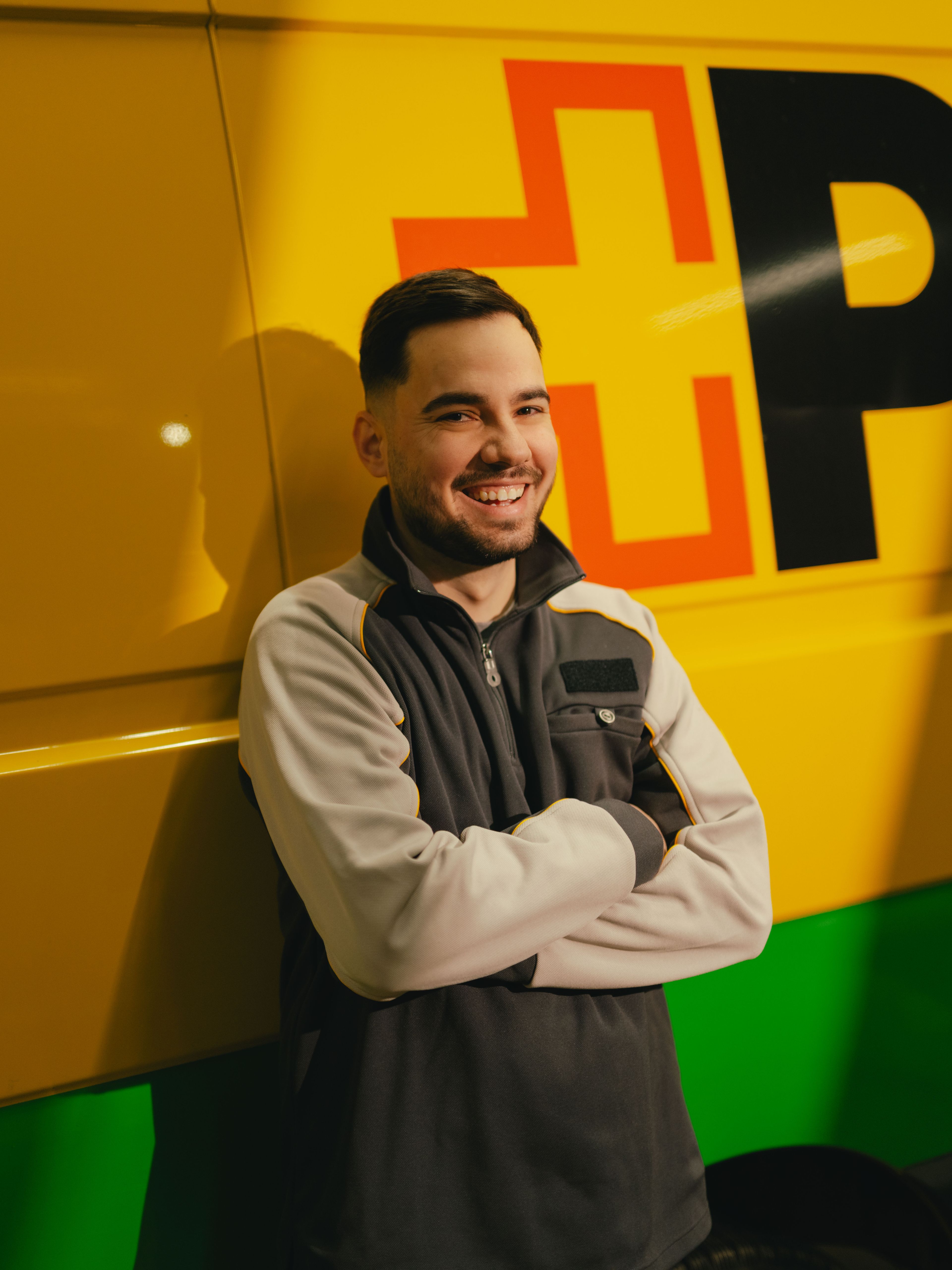 Eine Person steht mit verschränkten Armen vor einem gelb‑grünen Lieferfahrzeug mit grossen, schwarzen und roten Buchstaben auf der Fahrzeugseite.