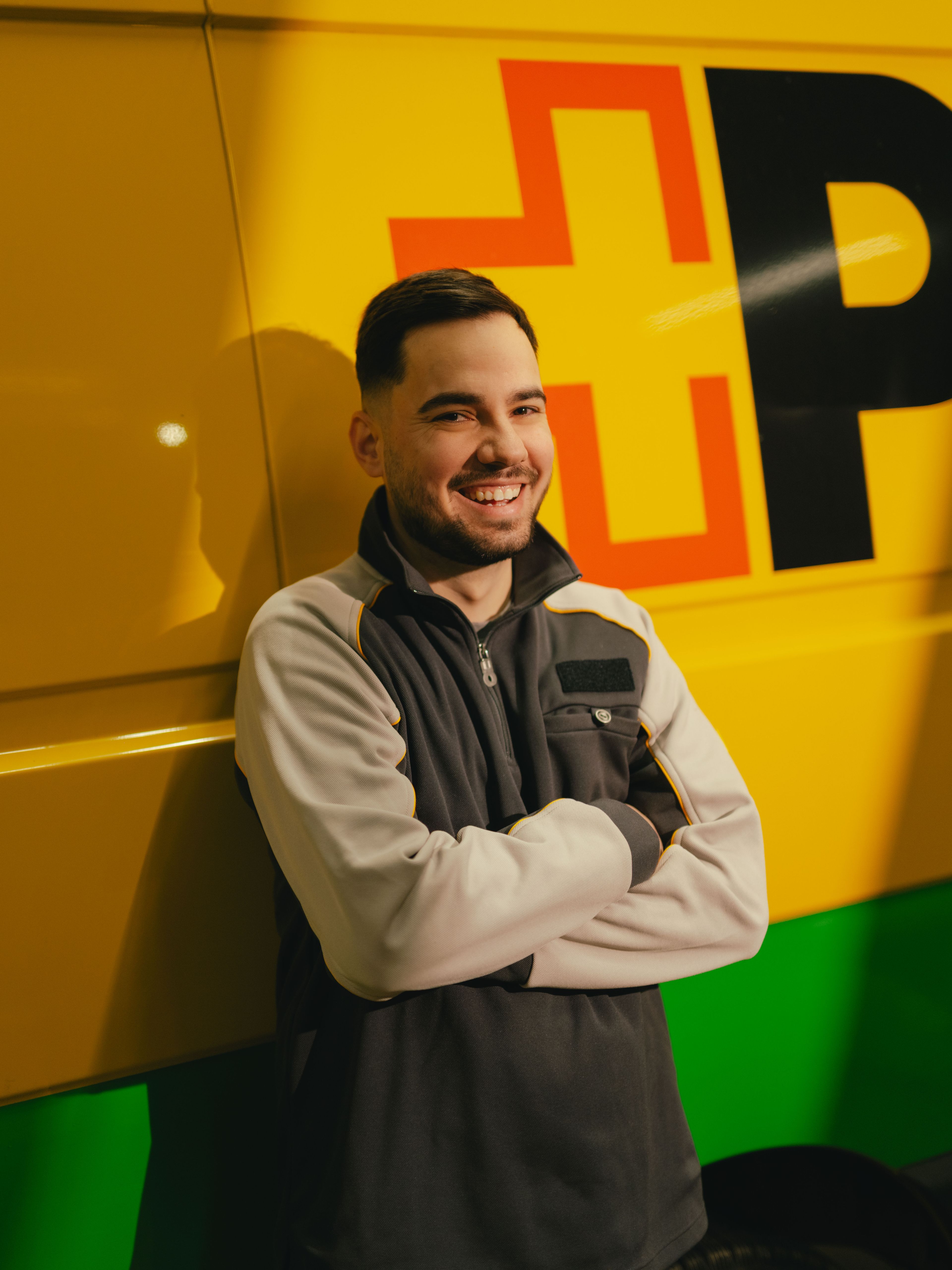 A person stands with arms crossed in front of a yellow and green delivery vehicle featuring large black and red lettering on its side.