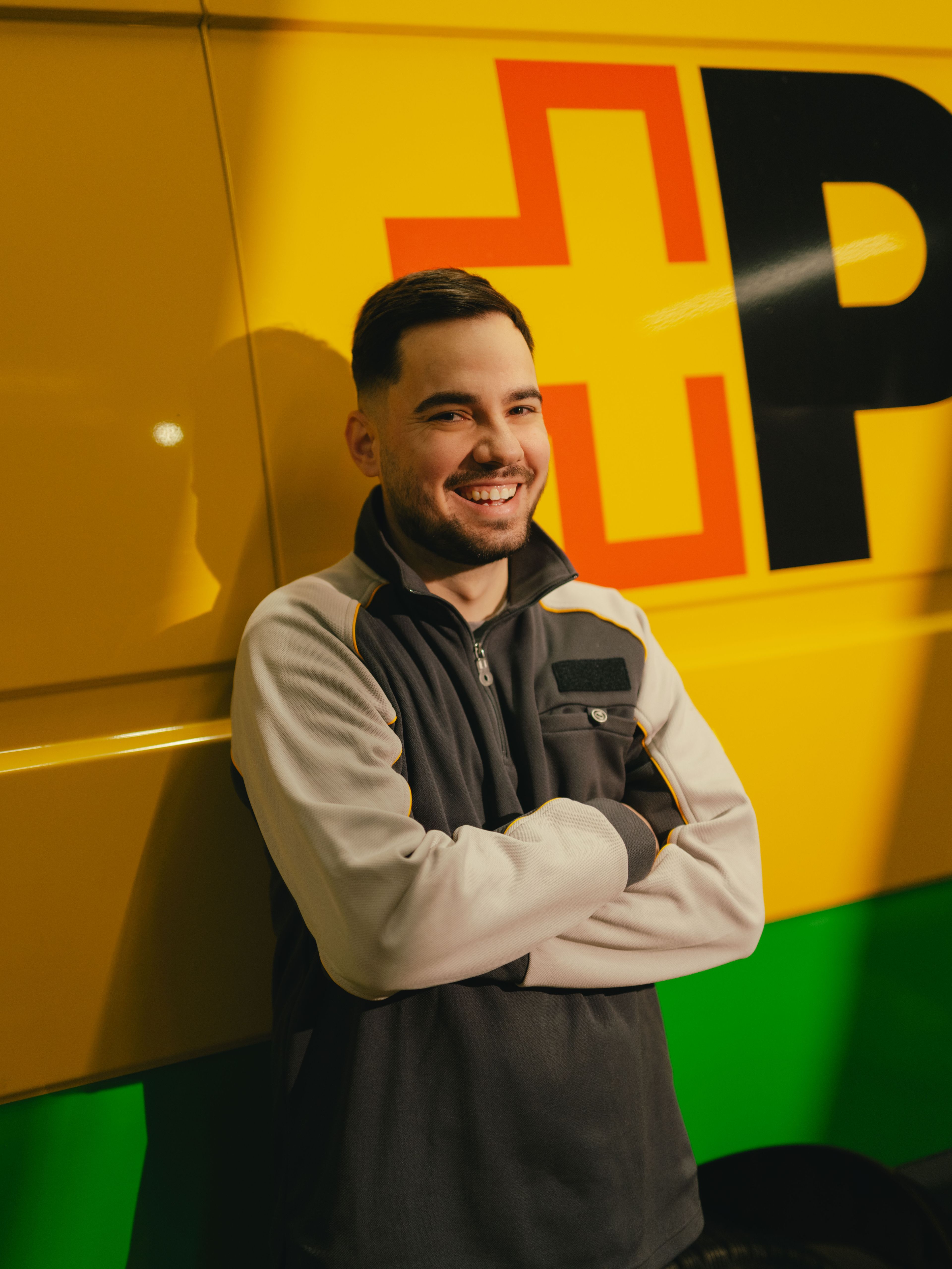 Une personne se tient les bras croisés devant un véhicule de livraison jaune et vert portant de grandes lettres noires et rouges sur le côté.