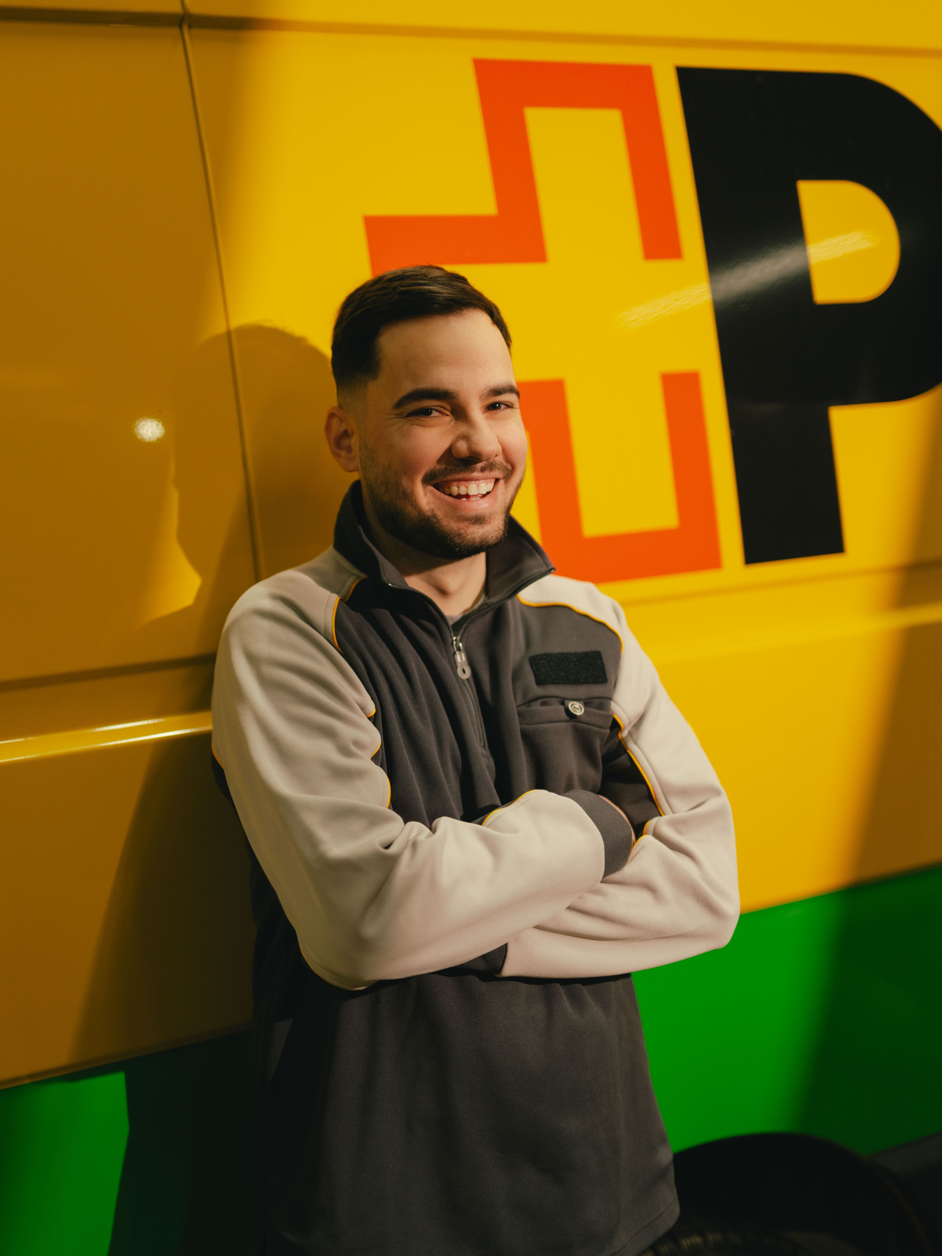 Une personne se tient les bras croisés devant un véhicule de livraison jaune et vert portant de grandes lettres noires et rouges sur le côté.