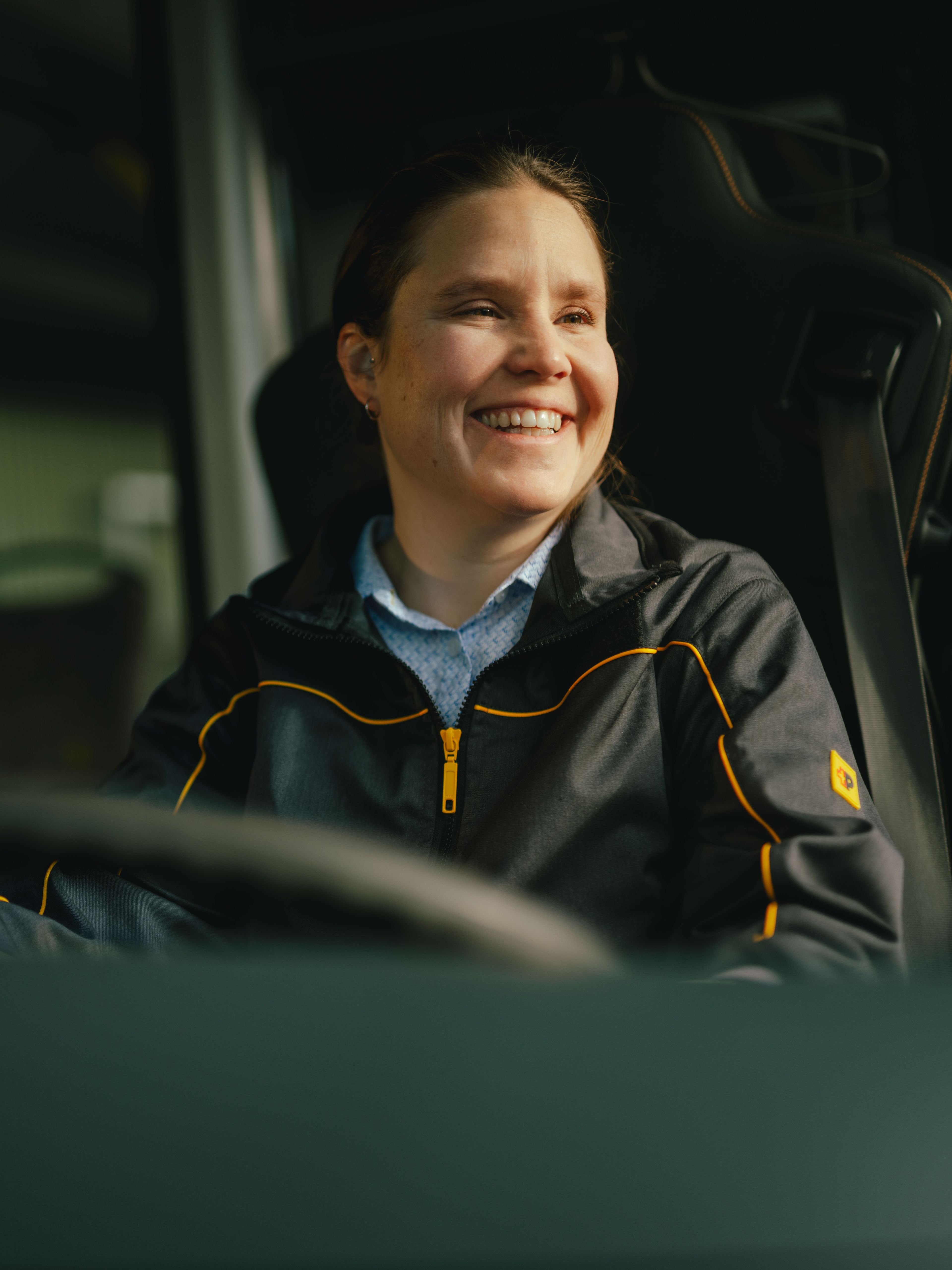 Eine Person sitzt am Steuer eines Fahrzeugs und trägt eine dunkle Jacke mit gelben Akzenten. Teile des Innenraums des Fahrzeugs, einschliesslich des Lenkrads und Haltegriffen, sind sichtbar.