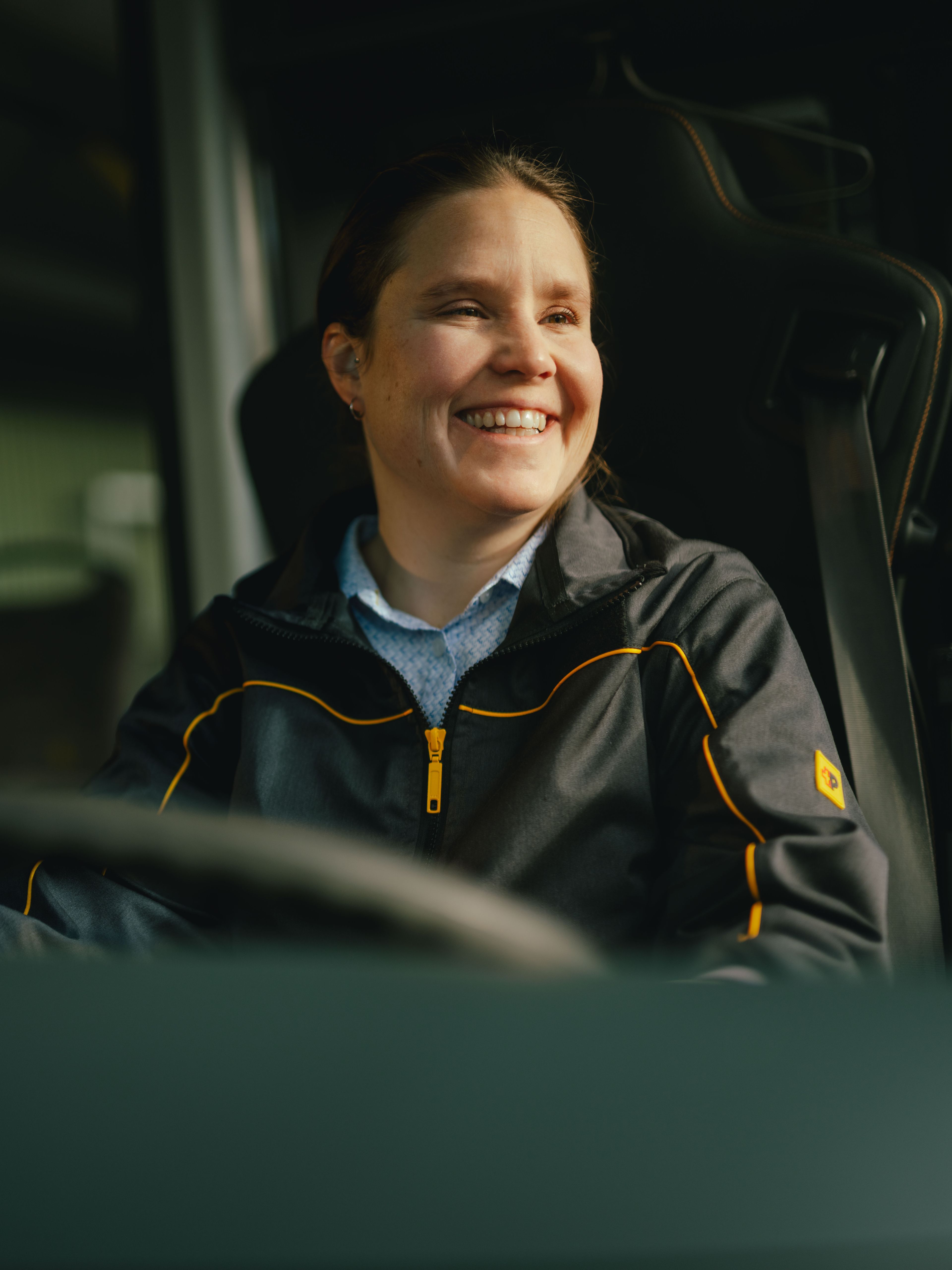 Une personne est assise au volant d’un véhicule, portant une veste foncée avec des accents jaunes. L’intérieur du véhicule, y compris le volant et des poignées, est visible.