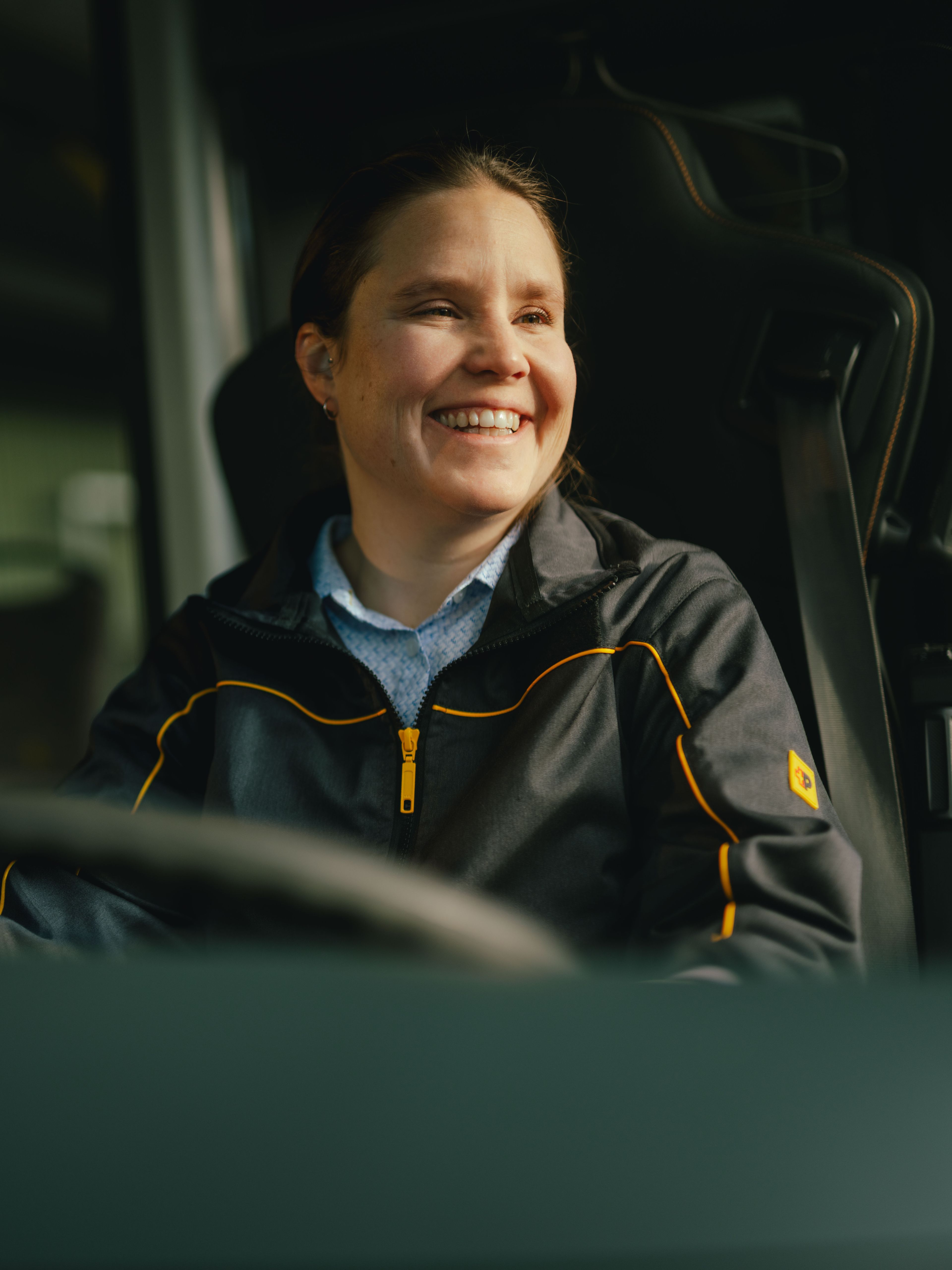 Una persona è seduta al volante di un veicolo e indossa una giacca scura con dettagli gialli. L’interno del veicolo, compreso il volante e alcune maniglie, è visibile.