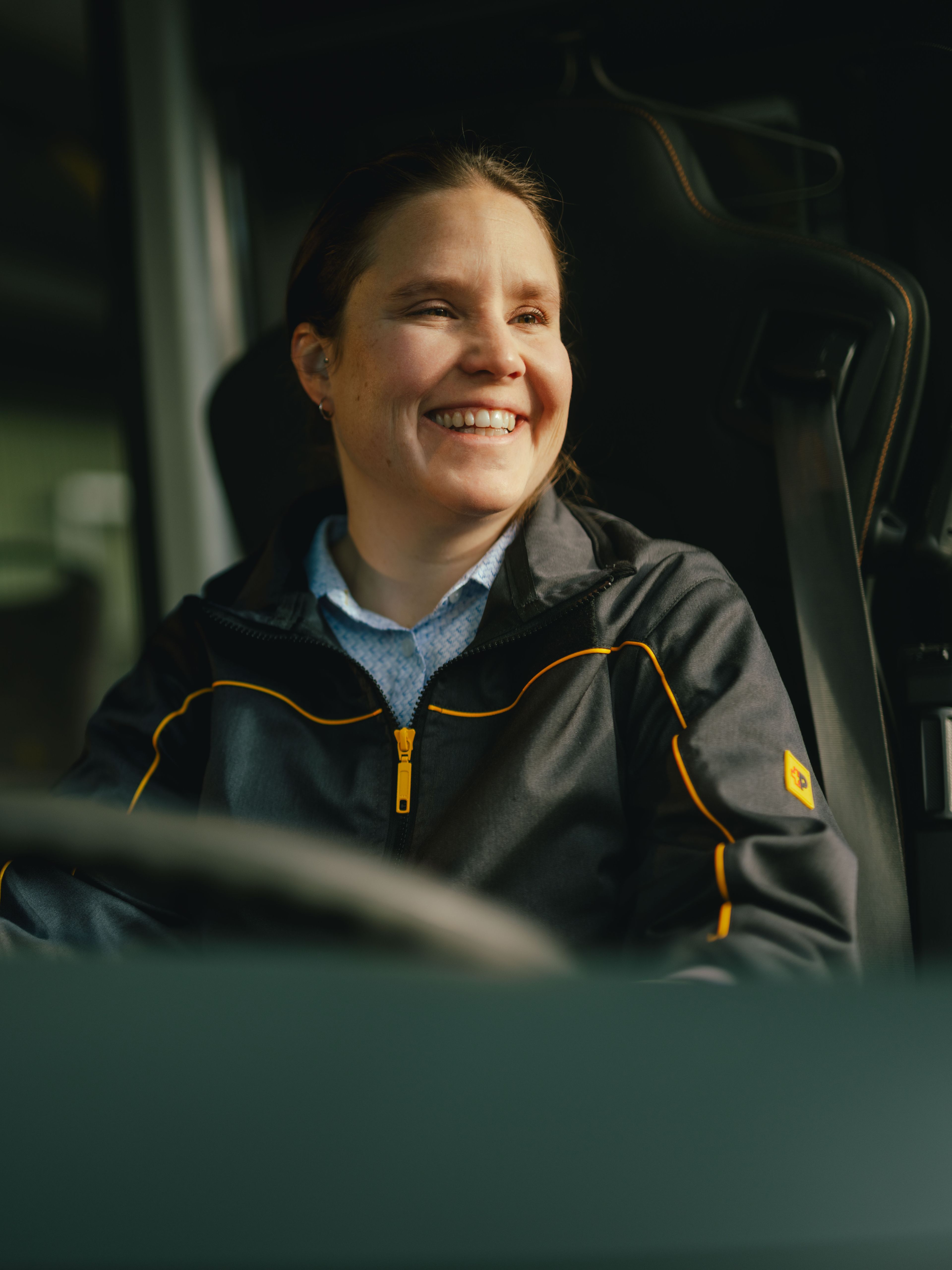 A person is seated in the driver’s seat of a vehicle, wearing a dark jacket with yellow accents. Parts of the vehicle’s interior, including the steering wheel and handrails, are visible.