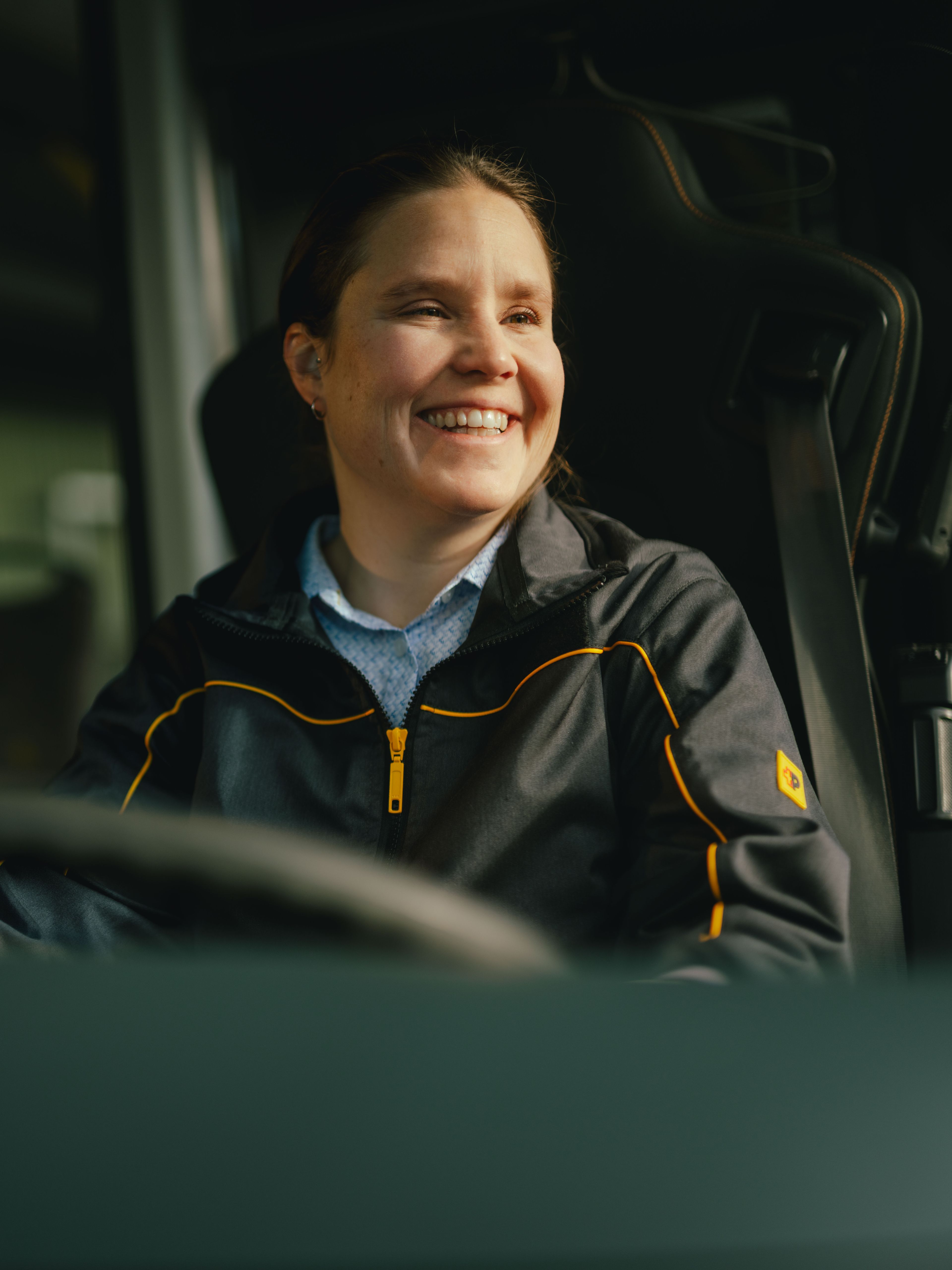 Una persona è seduta al volante di un veicolo e indossa una giacca scura con dettagli gialli. L’interno del veicolo, compreso il volante e alcune maniglie, è visibile.