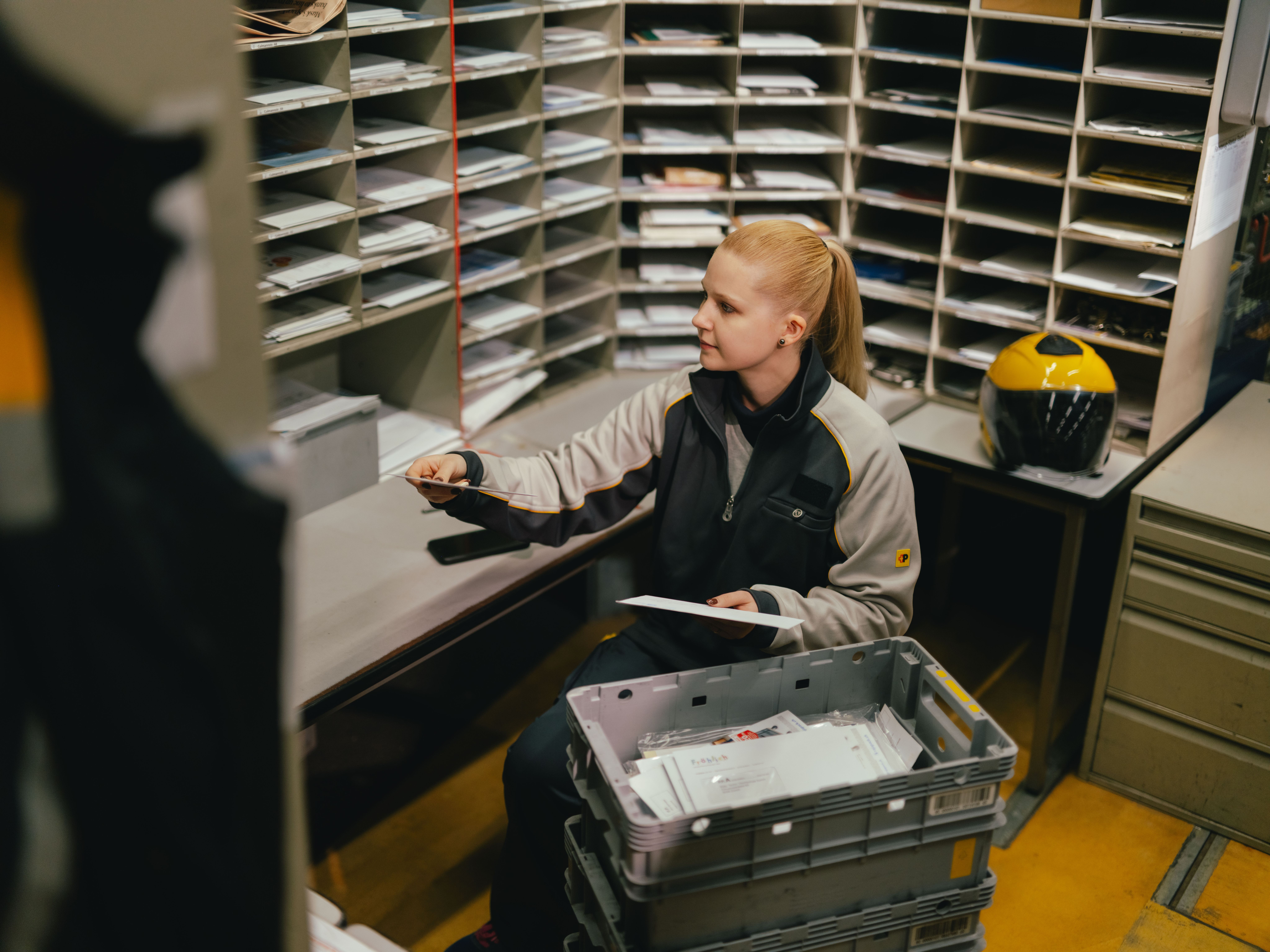 A person sits in a room filled with mail slots and is sorting letters. In front of the person is a grey plastic crate containing mail. Additional envelopes lie on the table, and a yellow helmet is positioned next to the workspace.