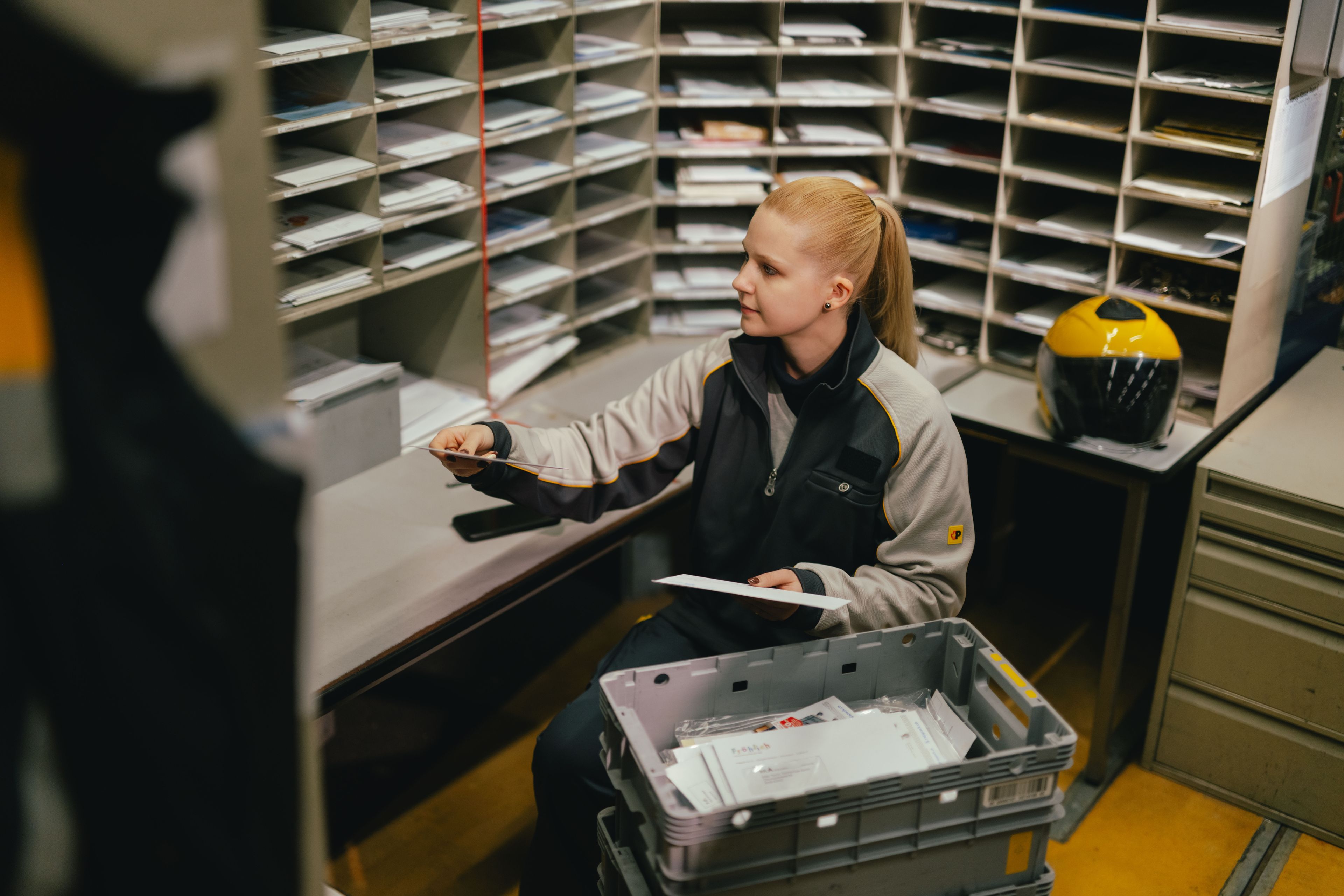 A person sits in a room filled with mail slots and is sorting letters. In front of the person is a grey plastic crate containing mail. Additional envelopes lie on the table, and a yellow helmet is positioned next to the workspace.